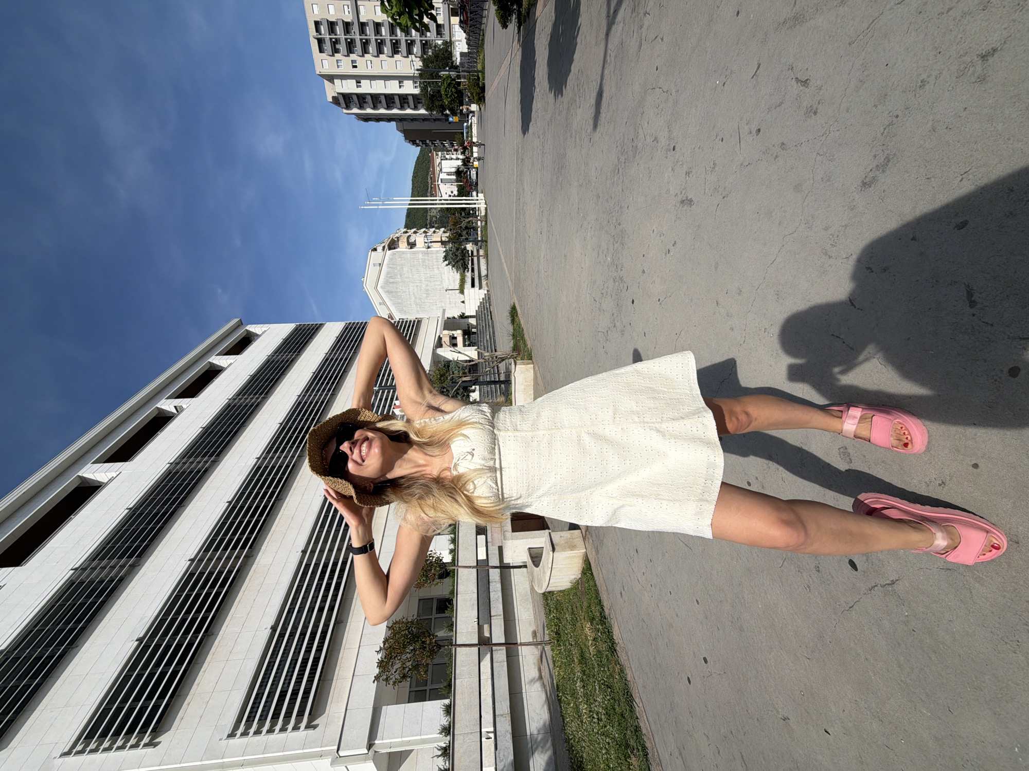 A young blonde woman with her hands on her head and wearing sunglasses and a straw hat stands on a paved path in a city square. She is wearing a short, white eyelet dress and bright pink platform sandals. In the background, a modern white building with horizontal windows is visible on the left, and residential buildings and trees line the right side of the square. The sky is clear and blue. The scene appears to be a sunny day in Budva, Montenegro.