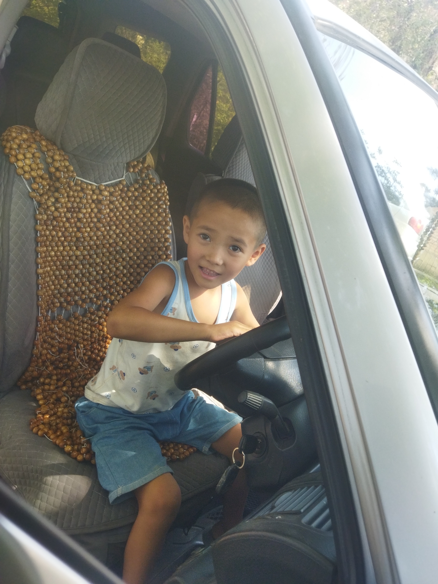 A young boy sits in the driver's seat of a car, looking towards the camera with a smile. He is wearing a white tank top with cartoon bear prints and blue denim shorts. His hands are on the steering wheel, and a key with a ring is visible in the ignition. The car seat behind him is covered with a beaded seat cover. The image appears to be taken on a sunny day, with light filtering into the car. The surrounding environment is blurred, but suggests an outdoor setting, possibly in a residential area.
