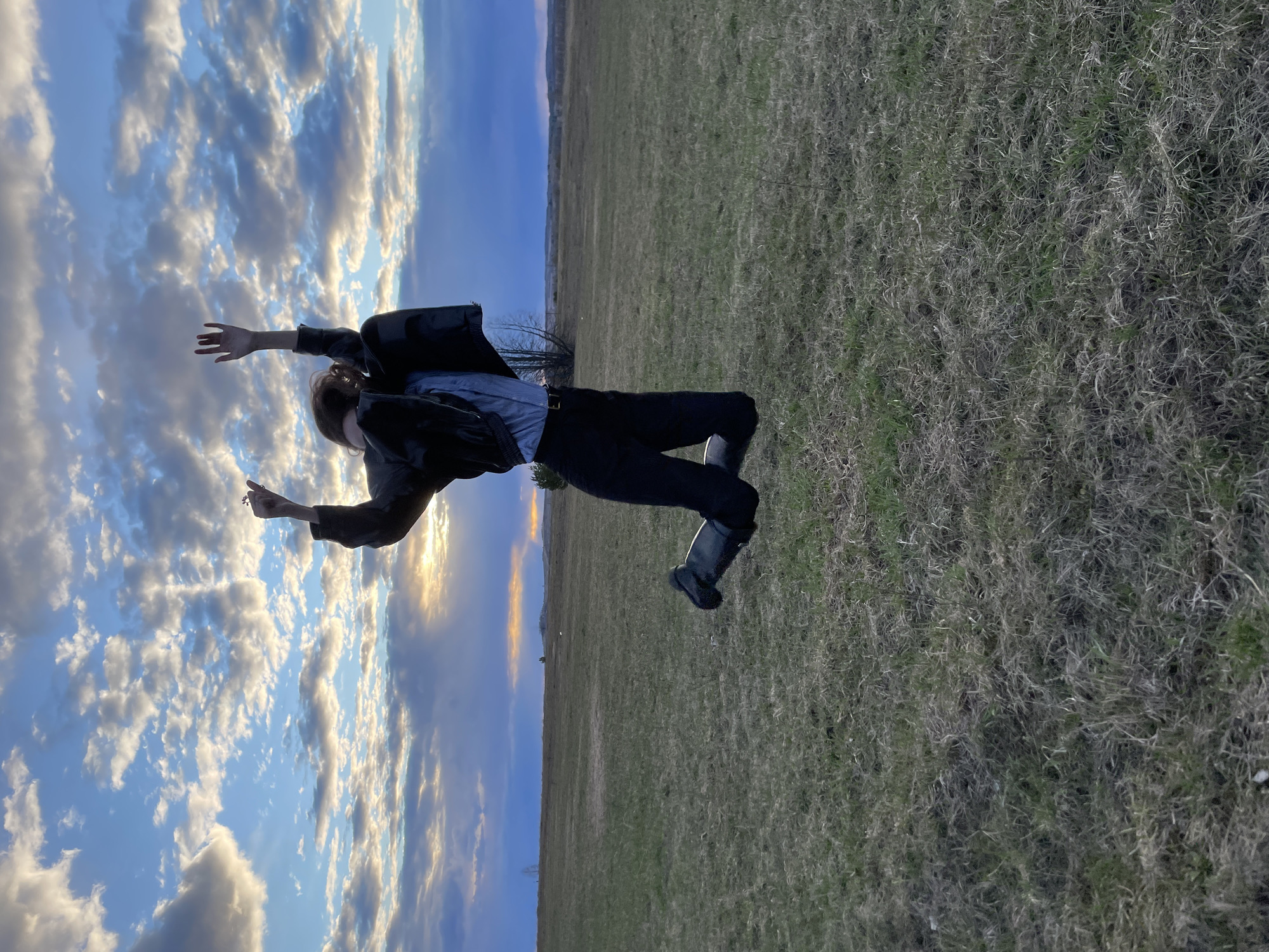 The image, oriented horizontally as if rotated 90 degrees clockwise, captures a person in an open field, seemingly suspended mid-air against a vast, cloudy sky. The individual, dressed in a black jacket, a light-colored collared shirt, dark pants, and dark boots, has their arms outstretched and legs bent, conveying a dynamic, weightless pose. Their hair appears to be light brown or reddish.
The background is dominated by an expansive sky on the left and an uncultivated landscape on the right. The sky is largely blue, filled with scattered white and grey cumulus and stratocumulus clouds. Patches of these clouds are illuminated by the warm, golden light of a setting or rising sun, indicating late afternoon or early evening. The ground, which appears vertically on the right side of the frame, is a large, open field covered with dry, sparse grass, interspersed with greener patches. A faint tree line is visible on the distant horizon.
The scene evokes a sense of freedom, playfulness, or an unusual perspective. No text is visible in the image. This outdoor scene is situated in Kueda, Russia.