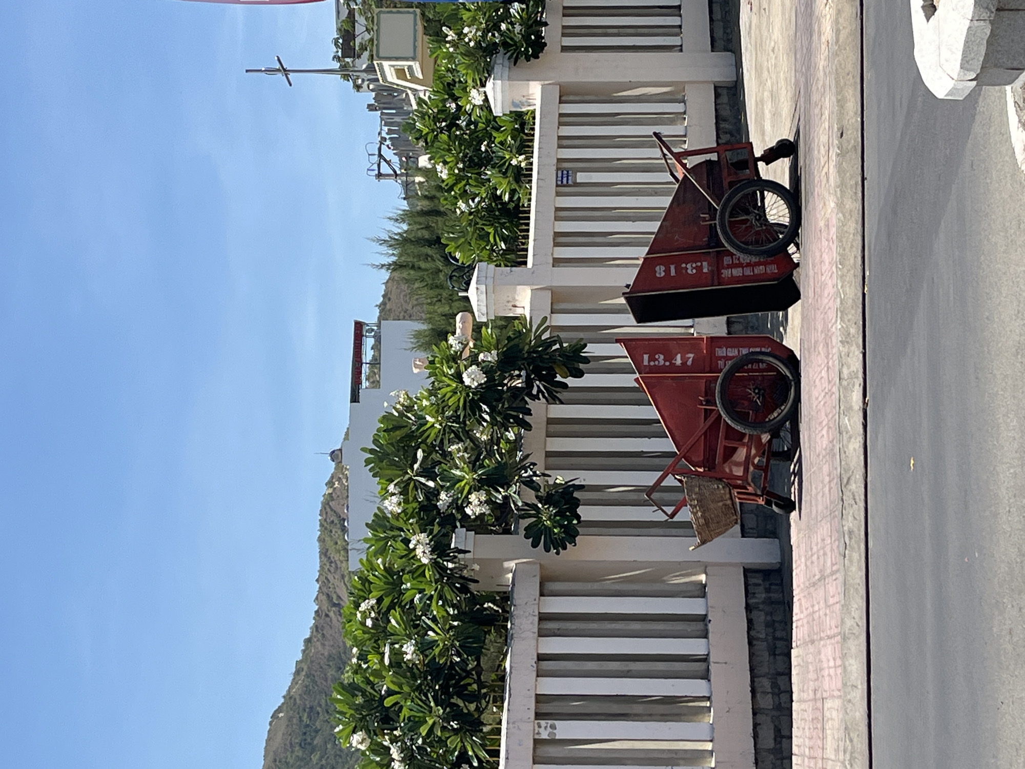 The image depicts a street scene in Bac Nha Trang Ward, Vietnam. Two red trash carts with black bins, labeled with numbers "1.3.47" and "81.18," are parked on the pavement next to a white fence. White flowering trees and green foliage line the fence. In the background, there are buildings and a hill under a clear blue sky.
