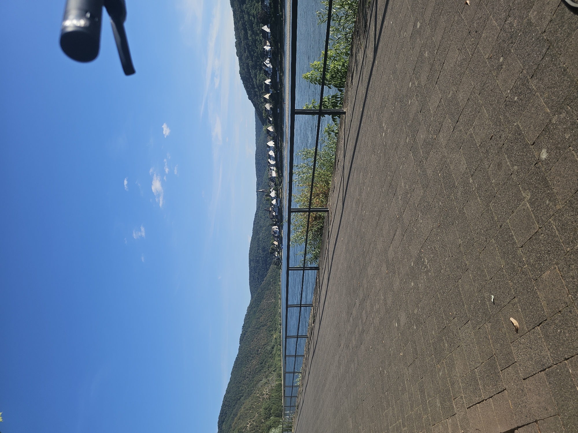 The image depicts a scenic view of the Rhine River in Braubach, Germany, on a clear, sunny day. A paved walkway runs along the foreground, bordered by a black metal railing. Beyond the railing, the wide, blue Rhine River stretches into the distance, reflecting the sky. On the opposite bank, a picturesque village with white-roofed houses is nestled at the base of lush, green, tree-covered hills. The sky is a vibrant, cloudless blue, with a few wispy clouds visible. In the upper right corner of the image, a blurry, black object, likely part of a camera or bicycle handlebar, is visible. The overall impression is one of tranquility and natural beauty.