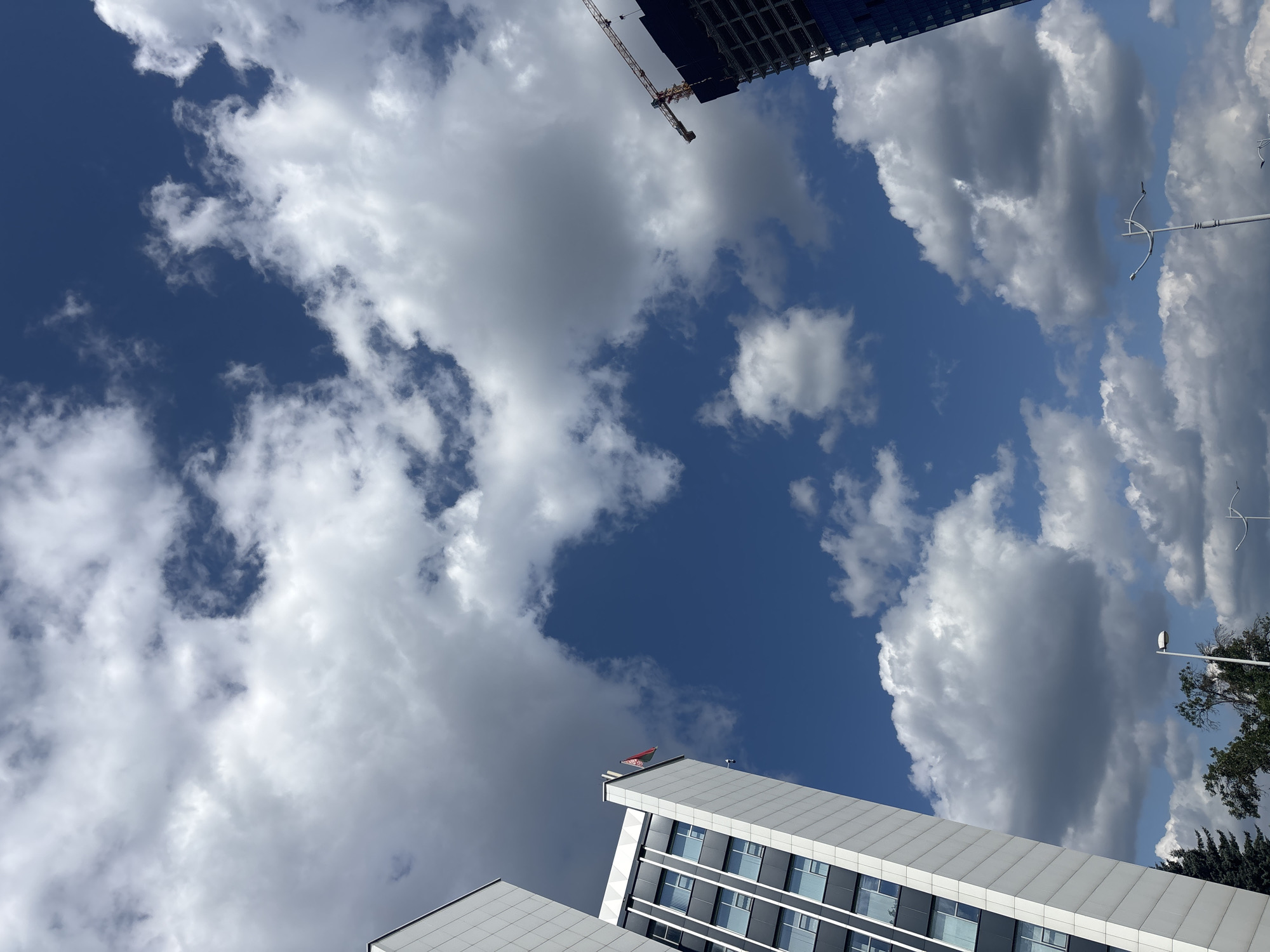 The image captures a skyward view of two modern buildings against a bright blue sky filled with fluffy white and grey clouds. On the left, a tall, sleek building with a white facade and dark-framed windows is prominently featured. A small red flag flies from the top of this building. On the right, a darker, unfinished skyscraper is visible, with a construction crane extending from its upper section. The scene suggests an urban environment, likely in Minsk, Belarus, given the presence of the national flag on the building. Trees and streetlights are visible at the bottom of the frame, further indicating an outdoor city setting. The overall atmosphere is one of daytime activity and ongoing development.