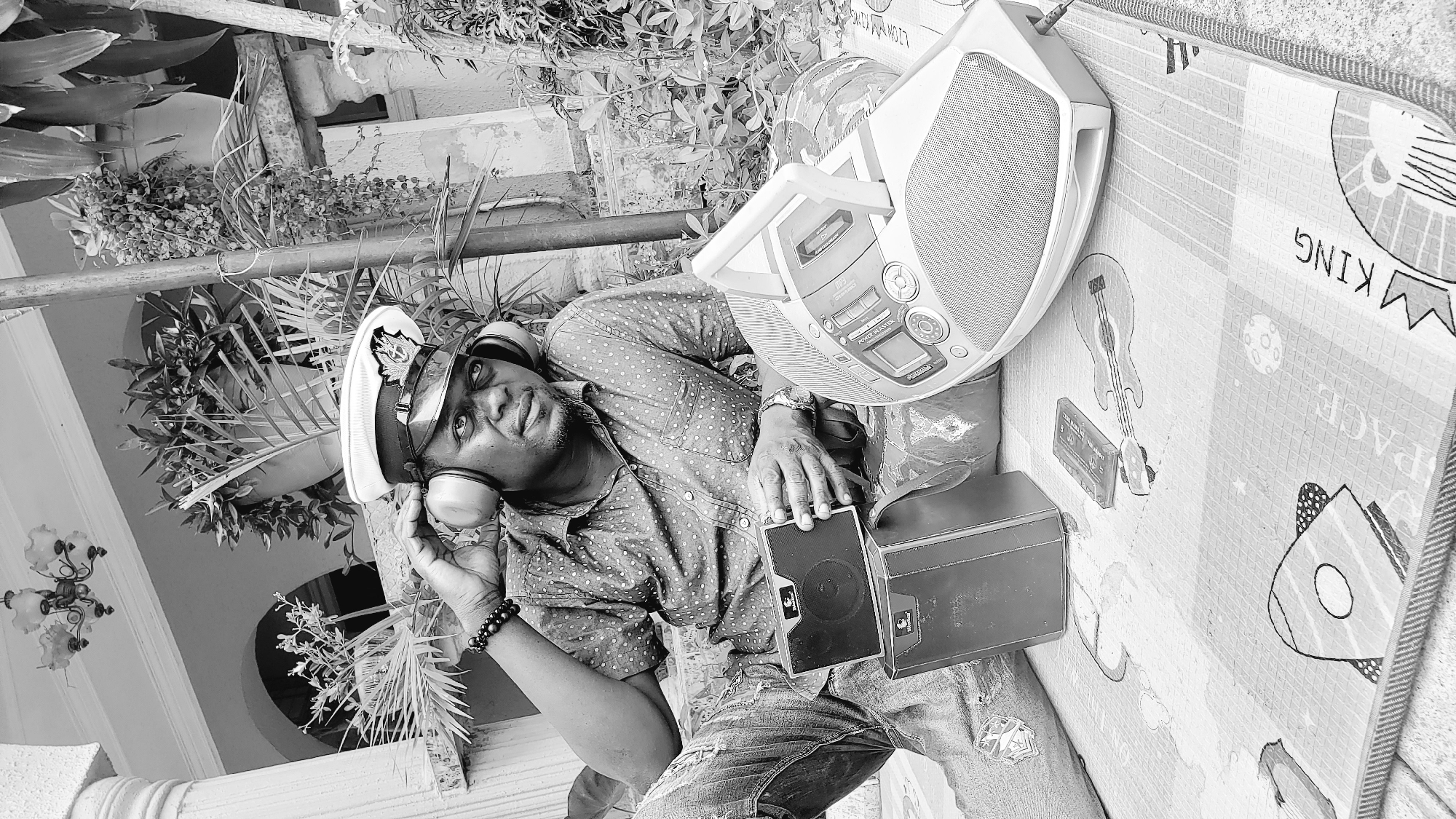 In Sabon Tasha, Nigeria, a Black man wearing a peaked cap and headphones is seated outdoors. He is dressed in a polka-dot shirt and jeans, with one hand resting on his headphones and the other holding a small speaker. In front of him are two stacked black boxes, a cassette tape, and a white boombox. The man is positioned on a patterned mat in a setting with lush greenery and architectural elements suggesting a veranda.