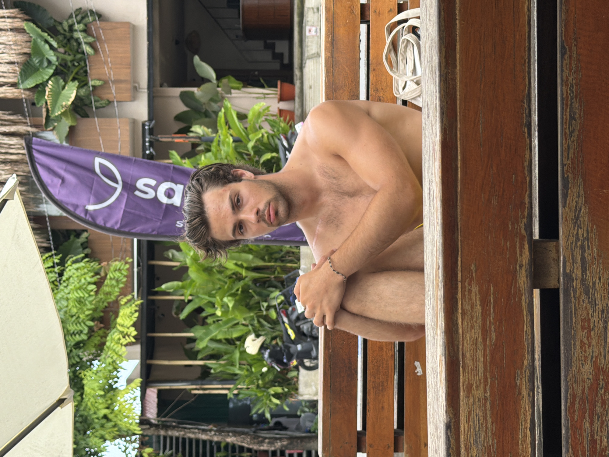 The image depicts a young man, shirtless and with dark hair, looking directly at the camera. He is in a crouched or seated position, with his left knee bent towards his chest and his right arm resting on his left leg. A thin, patterned bracelet is visible on his left wrist. He has a neutral to slightly pensive expression.
He is positioned beside a weathered wooden structure, likely a bench or barrier, which frames the right side of the image. A white strap or cord is loosely coiled and hanging on this wooden structure behind him. To his left, a large white umbrella or canopy is visible, partially obscuring the background.
The background features lush green tropical foliage, suggesting an outdoor setting in a warm climate. Faintly visible behind the greenery are what appear to be buildings or houses. In the mid-ground, a purple banner or flag is prominent, displaying a white leaf-like logo and the partial text "sa". Below this, two motorcycles or scooters are parked.
The scene appears to be a casual outdoor area, possibly a street, cafe, or beachfront spot. The lighting indicates it is daytime. Given the context of General Luna, Philippines, the tropical vegetation and casual outdoor setup are consistent with the location.