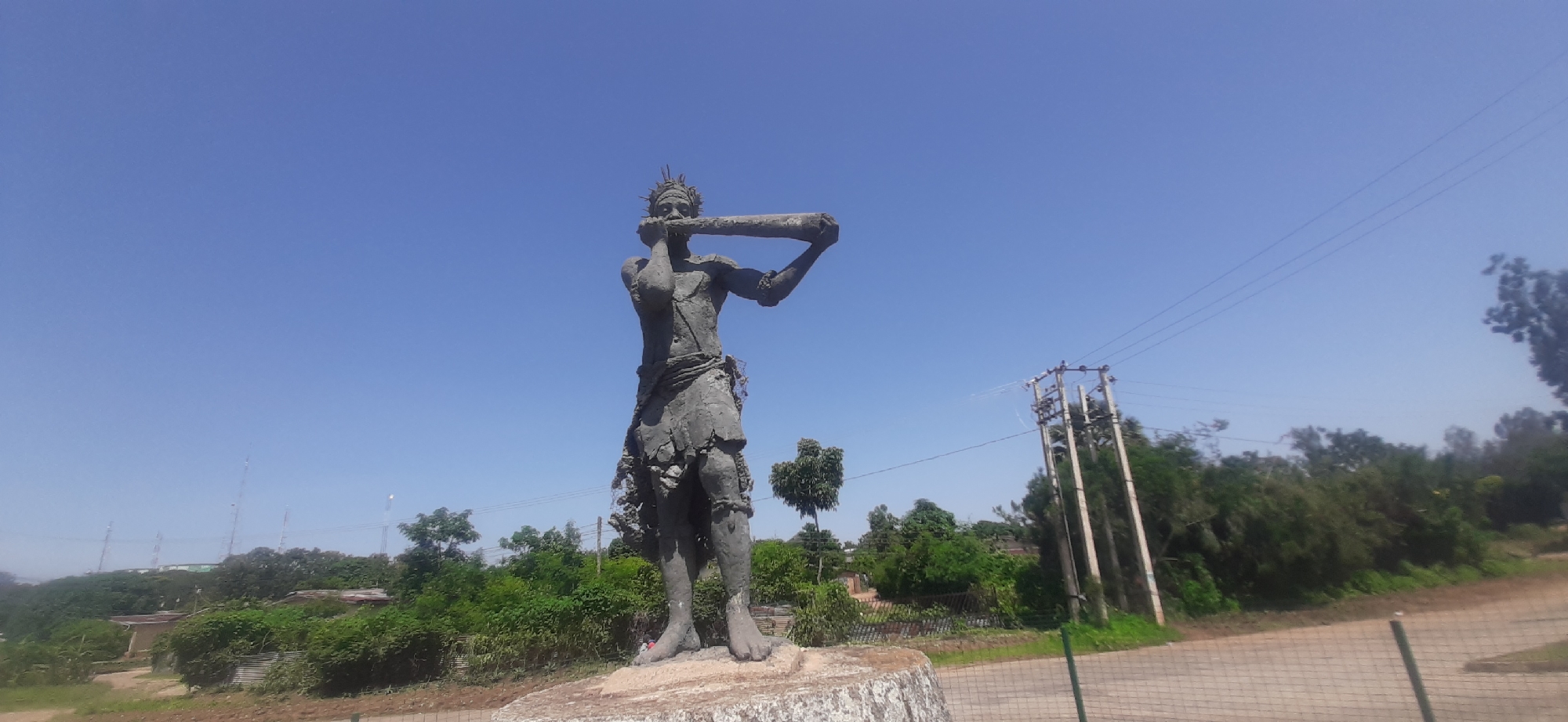 This is a full, wide-angle shot of a statue of a man playing a horn, positioned outdoors in a clear, blue sky. The statue is dark grey and appears to be made of rough-hewn material, possibly stone or cement. It depicts a muscular male figure with a headdress and what appears to be a skirt or loincloth. His arms are raised, holding a long, cylindrical object to his mouth, resembling a horn or a large trumpet. The statue stands on a rough, light-colored circular base.
The background consists of lush green vegetation, including trees and bushes, with some corrugated iron structures and a fence visible on the right. Power lines and utility poles are present in the mid-ground and background, indicating a developed area. In the far background, several tall communication towers are visible on a hill. The lighting suggests it is daytime, likely sunny, with no visible shadows cast by the statue itself due to the wide-angle lens and perspective.
While the exact location within Jos, Nigeria is not explicitly stated in the image, the presence of communication towers and the general landscape are consistent with such an environment. The scene is static, with no human activity depicted other than the figure represented by the statue.