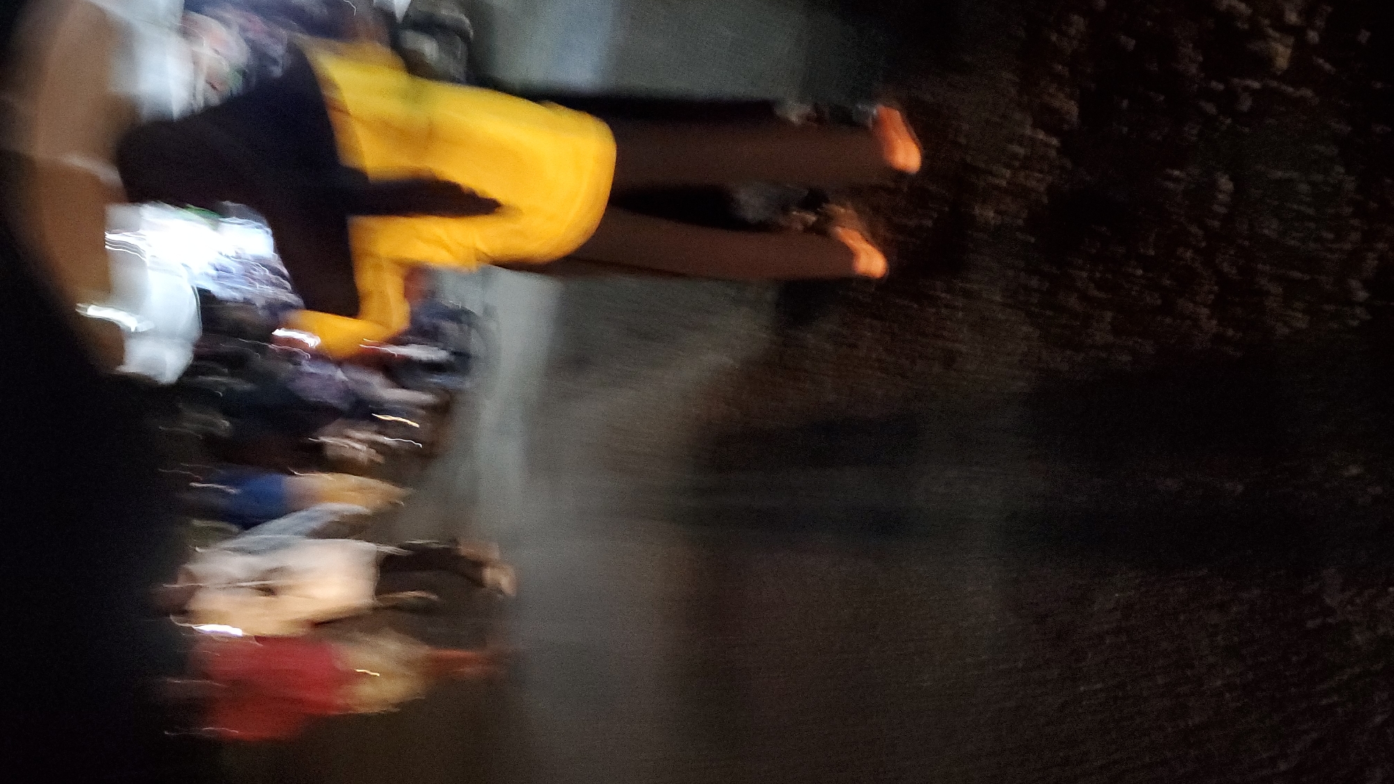 A blurry, low-light, outdoor nighttime scene in Jalingo, Nigeria, shows several individuals, mostly out of focus. In the foreground, a person wearing a bright yellow jacket and dark pants is visible from the waist down, with orange shoes. To the left of this person, other figures are seen in motion. One person appears to be wearing a white garment, and another is wearing a red garment. In the background, more indistinct figures and possibly some structures or tents are present. The ground appears wet or reflective, suggesting recent rain or standing water. The overall impression is of a gathering or event taking place after dark in an urban setting.