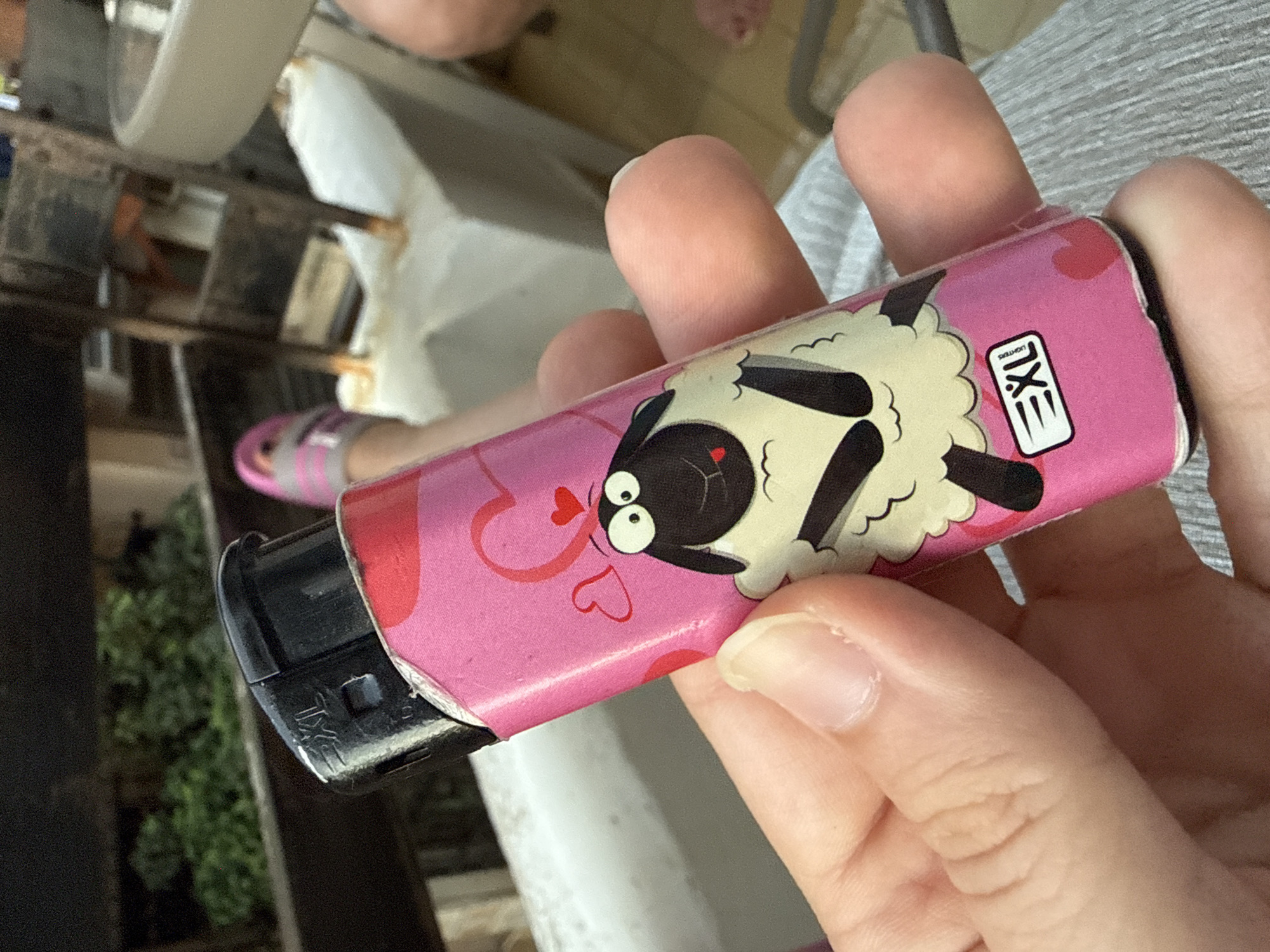A person's hand holds a pink disposable lighter decorated with cartoon sheep and hearts. The lighter has a black cap on one end. The background is blurred but suggests an indoor setting, possibly a home, with some furniture and possibly plants visible. The overall impression is casual. The visible text on the lighter reads "EXL LIGHTERS." The image is taken in Sozopol, Bulgaria.