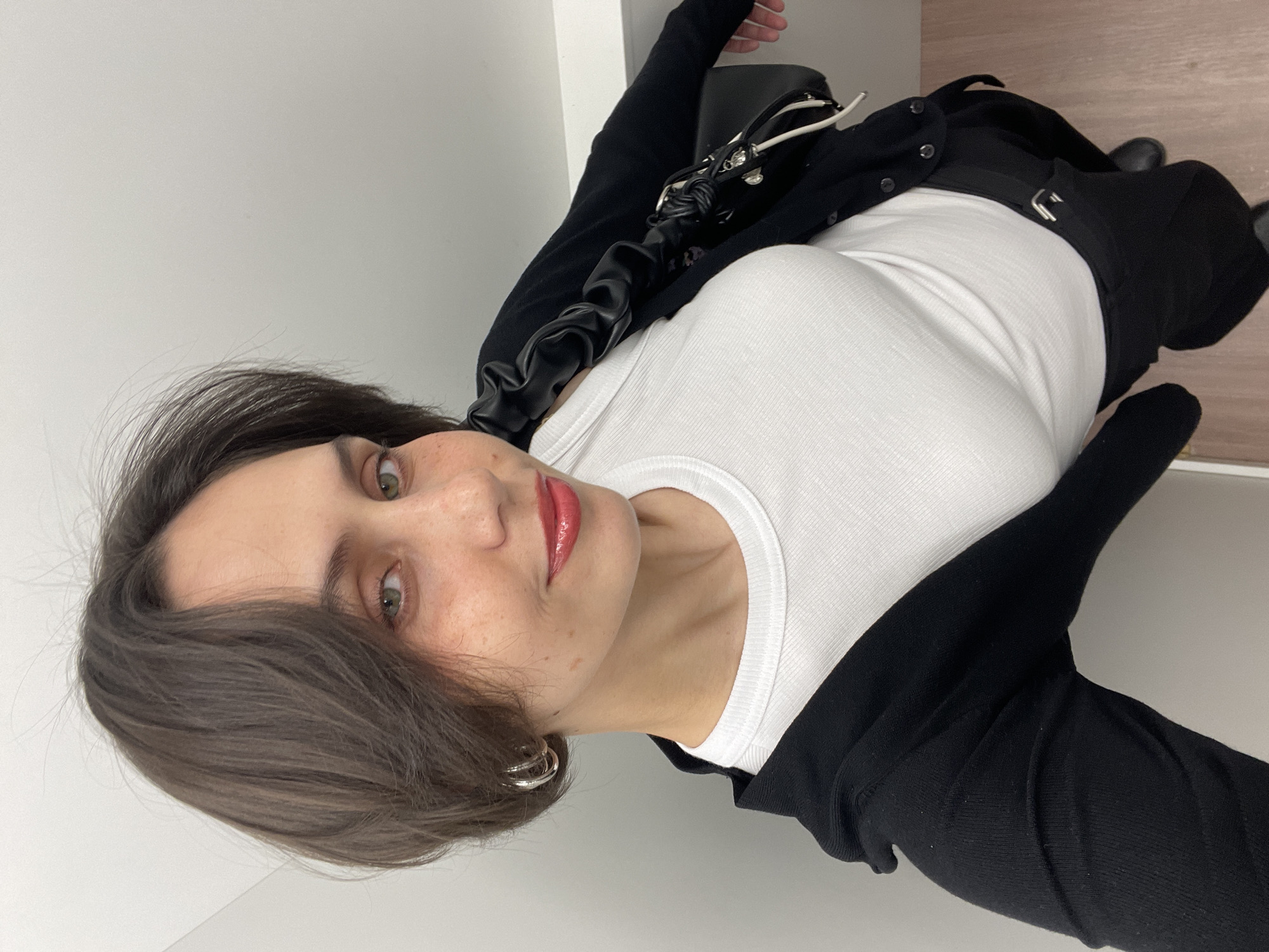 A woman is posing for a photograph. She is wearing a white ribbed tank top, a black cardigan, black pants with a silver buckle, and black shoes. She also has a black purse with a white strap. The woman has short brown hair, green eyes, and is wearing red lipstick. She is wearing silver hoop earrings. The background is a light gray wall and a light brown floor. The location in the city of Izhevsk, Russia is unknown.