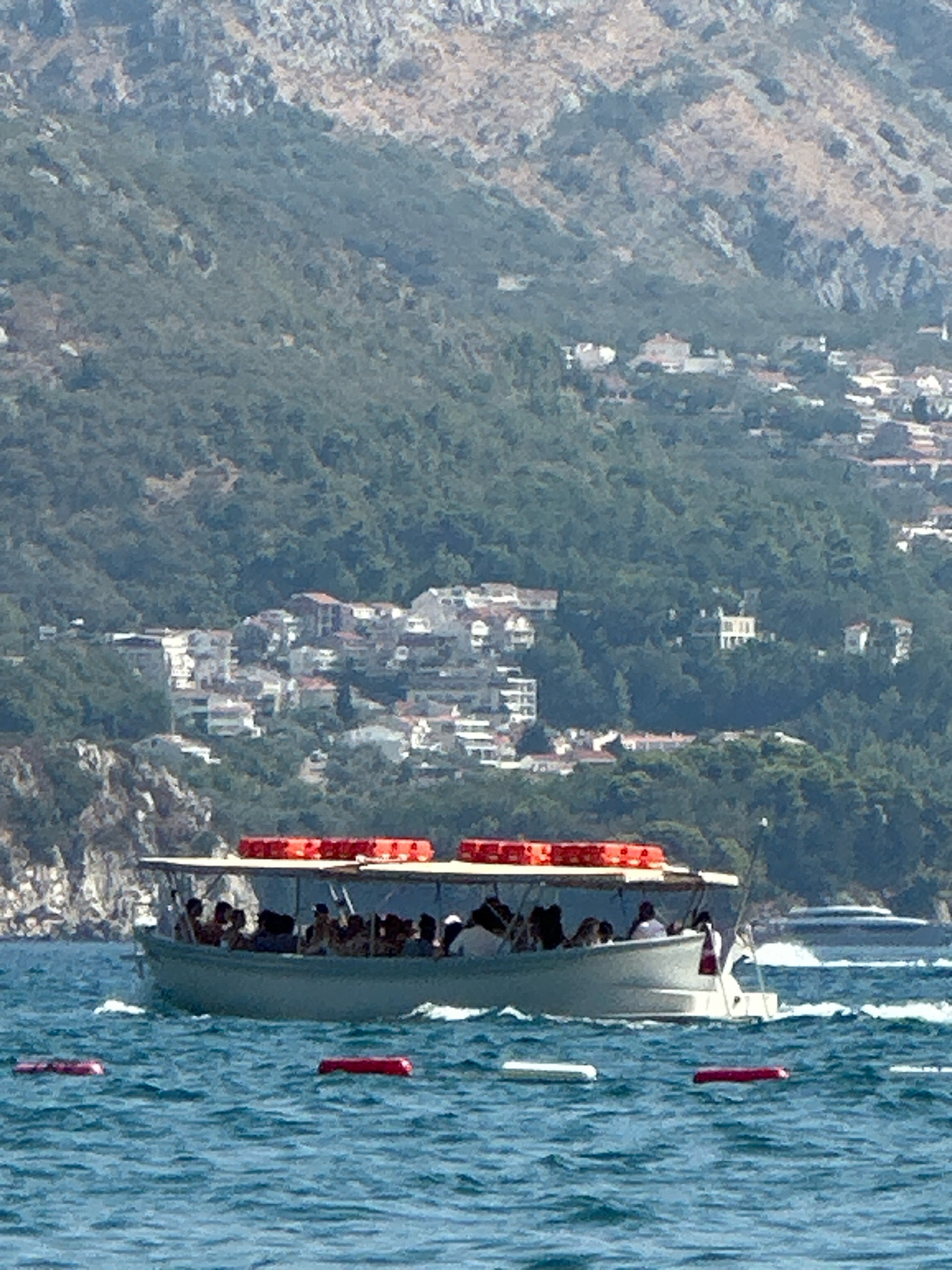 A medium shot captures a light-blue tourist boat filled with passengers on choppy, blue water. The boat has a white canopy with several orange life vests neatly stacked on top. The passengers are mostly silhouetted, with a few distinct figures visible. In the background, a densely populated town with white and red-roofed buildings clings to a lush, green hillside. The hillside is characterized by rocky outcrops and dense foliage, suggesting a Mediterranean coastal environment. Further in the distance, more distant, hazy buildings can be seen on the higher slopes of the mountain. The scene is under bright daylight. The area is identified as Boreti, Montenegro.