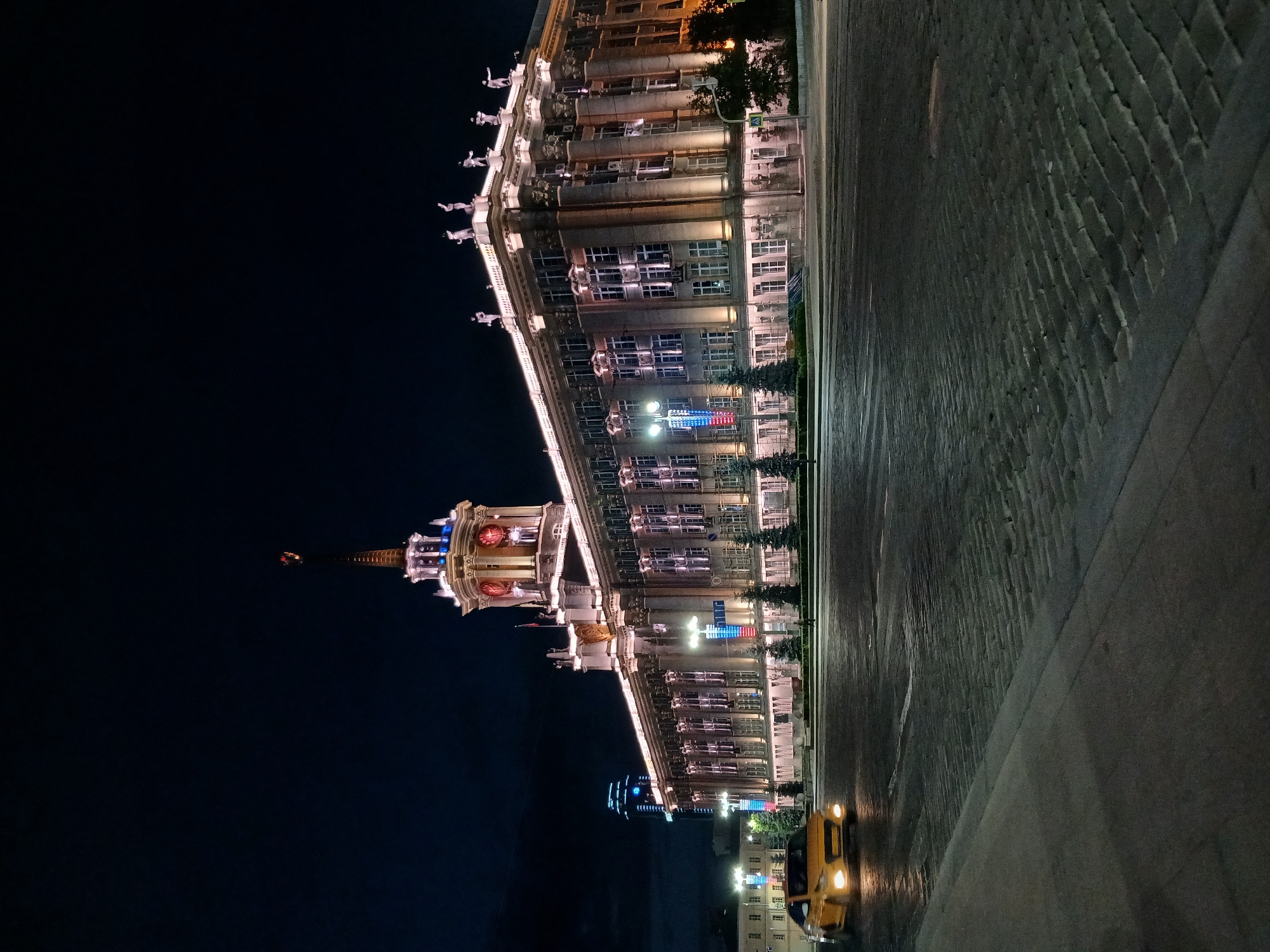 The image depicts the Yekaterinburg City Administration building at night, located in Yekaterinburg, Russia. The imposing Stalinist Empire style building is illuminated, with lights highlighting its architectural features, including statues adorning the rooftop. A clock tower with red clock faces is a prominent element of the structure. In the foreground, a yellow car is driving on a cobblestone street. The sky is dark, indicating nighttime. The scene is devoid of significant human activity, with only a few indistinct figures visible in the distance. The overall atmosphere is one of urban tranquility and grandeur.