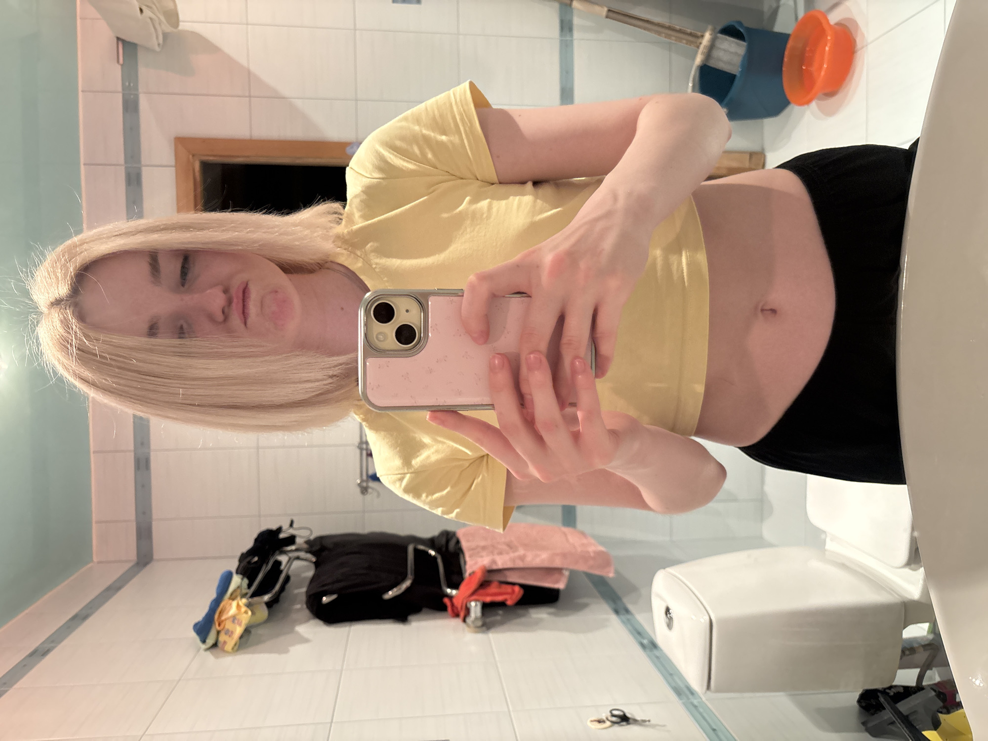 In a bathroom in Sinyavino, Russia, a young Caucasian woman with blonde hair and fair skin takes a mirror selfie. She is wearing a yellow crop top and black pants, with her midriff exposed. She is holding a pink phone with a floral pattern in her hands, and she has a pouty expression on her face. The bathroom is tiled with white square tiles, and there is a toilet, a sink, and a towel rack with several towels hanging on it. There are also some cleaning supplies visible in the background, including a bucket and a mop.