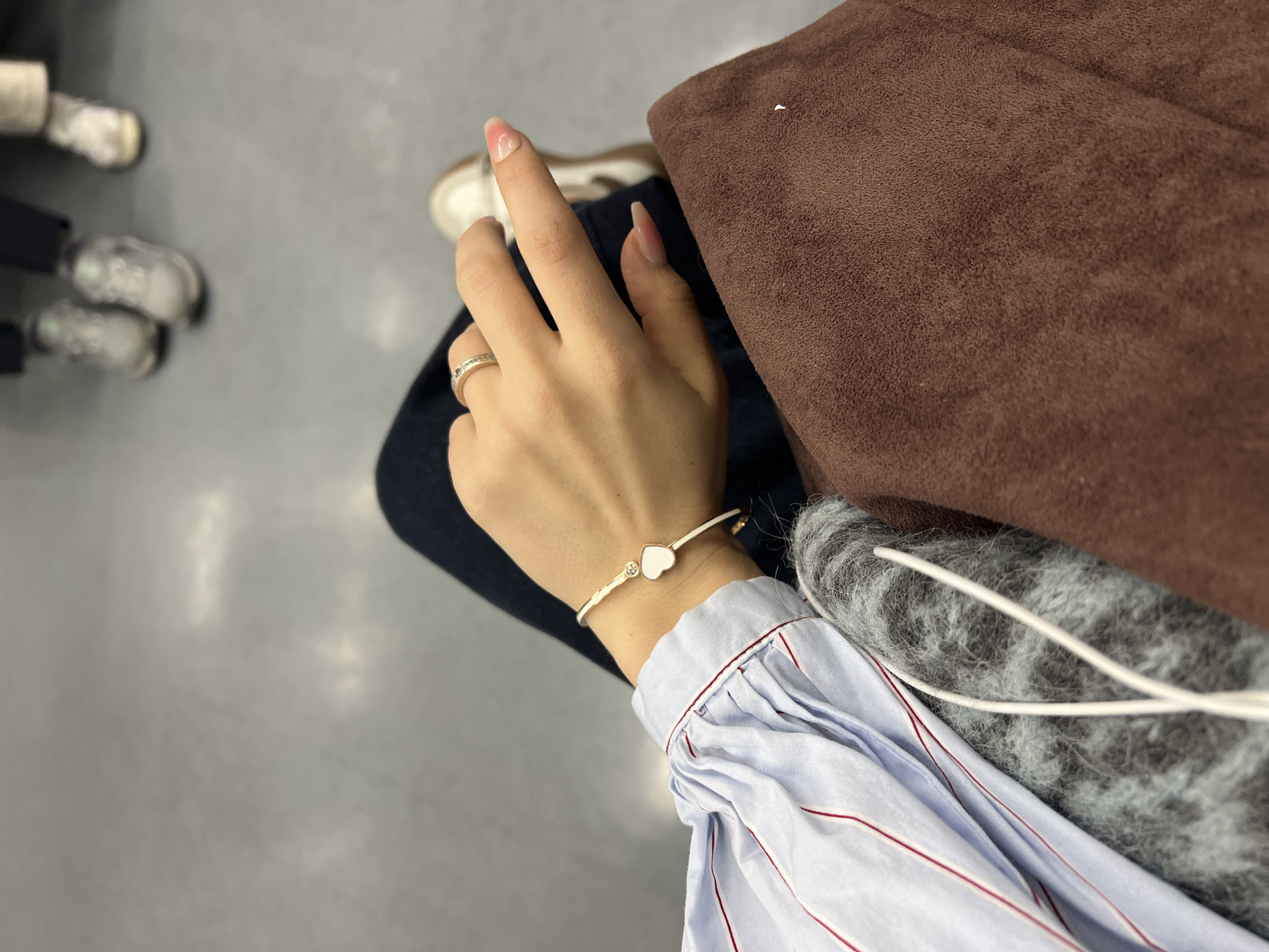 A person is seated indoors, likely in a public space. The individual is wearing dark-colored jeans, a light blue and white striped long-sleeved shirt, and white sneakers with brown accents. They are wearing a white bracelet with a small heart charm on their left wrist, and a ring on their left ring finger. Beside them sits a brown suede bag and a gray fuzzy shawl or sweater. Two pairs of feet and shoes belonging to other people are visible in the blurred background. The location is unknown, and there is no indication this photograph was taken in Seoul, South Korea.