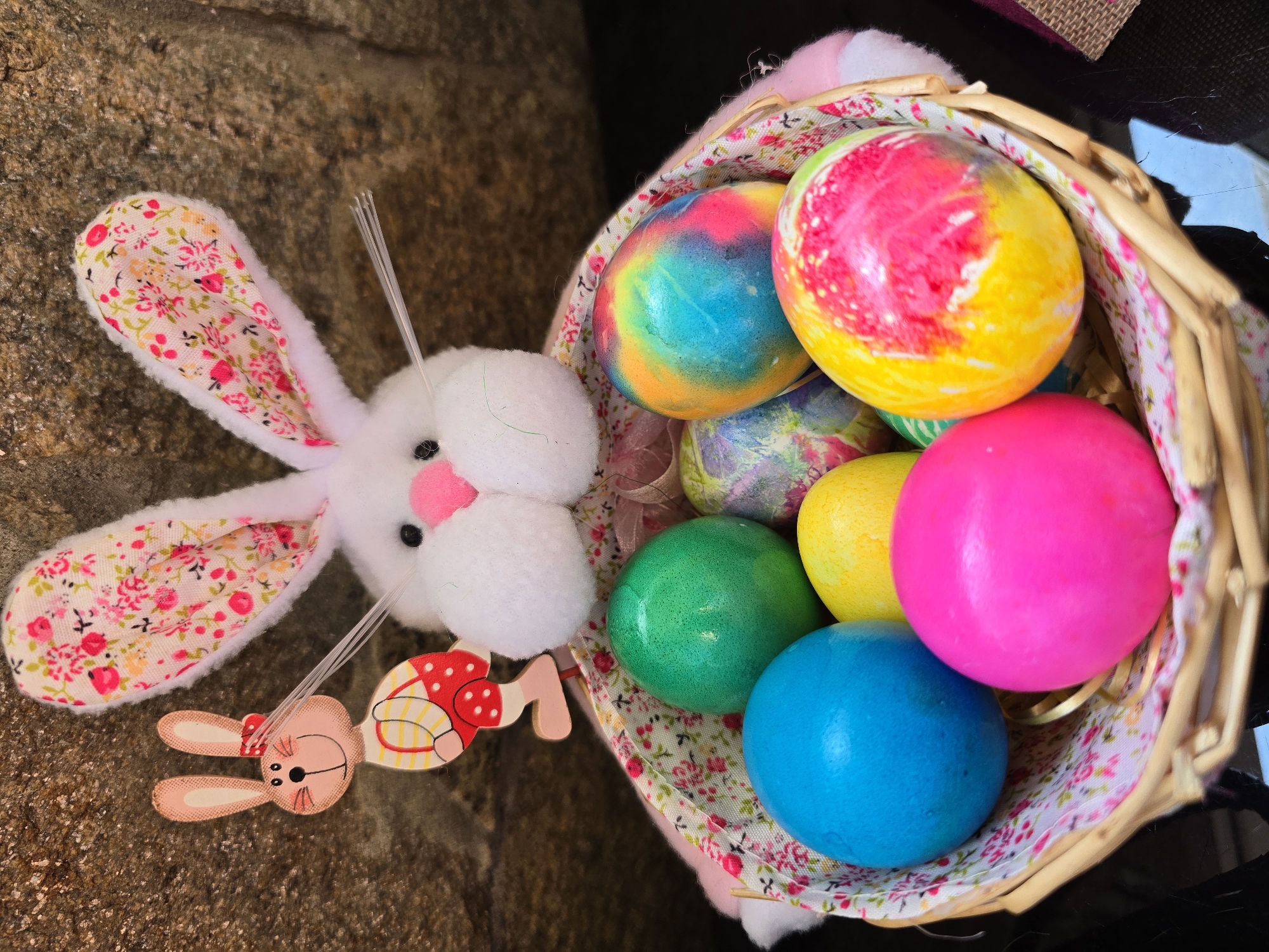 A basket overflowing with brightly colored Easter eggs sits next to a plush, white bunny with floral print ears and a wooden bunny charm, all resting on a stone surface. The egg colors include blue, green, yellow, pink, and a tie-dye pattern. The scene is outside. There is no evidence to determine the location is Burgas, Bulgaria.