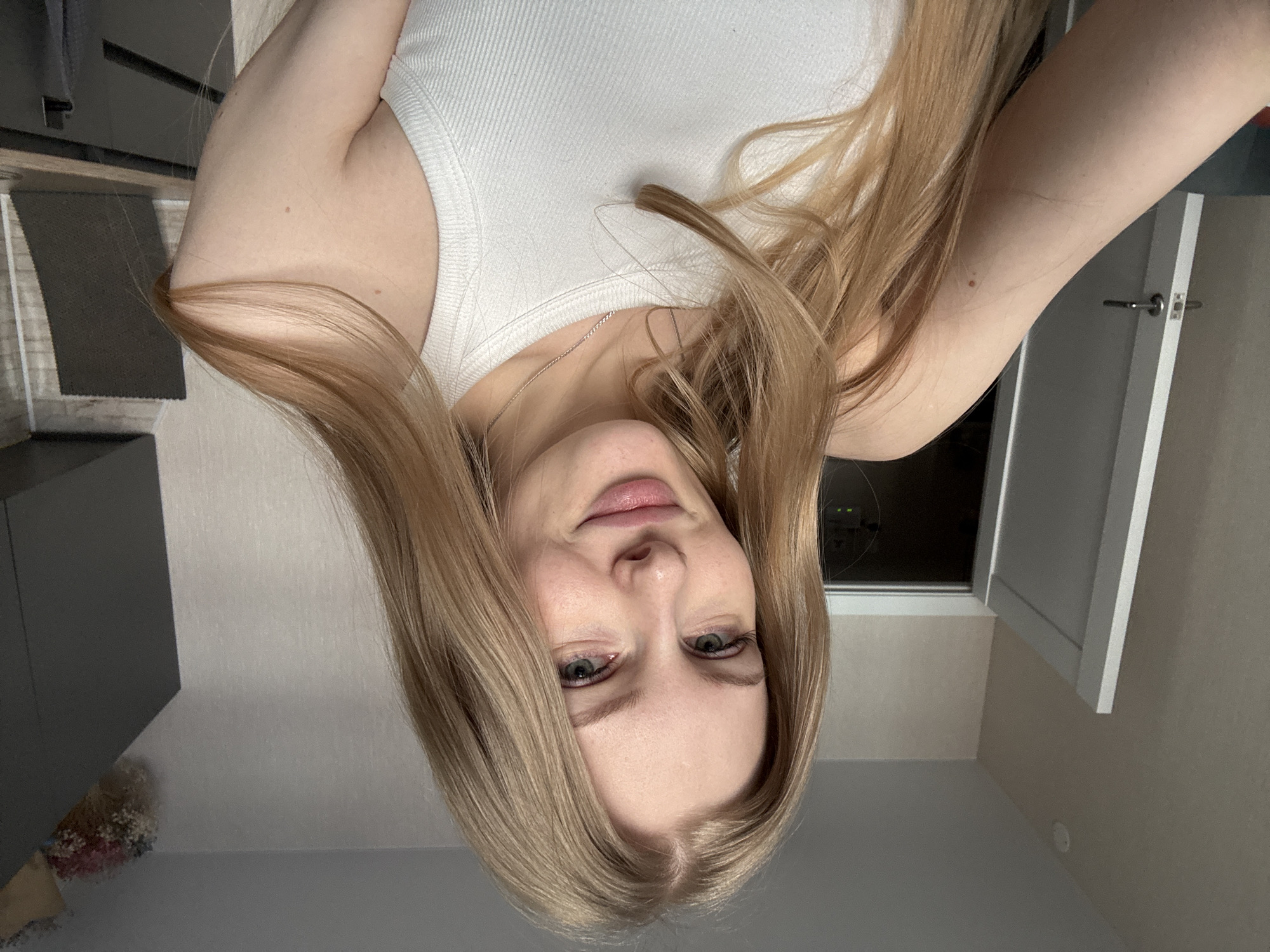 A selfie taken indoors, showing a woman with blonde hair wearing a white tank top. The location is likely an apartment based on the interior elements visible. The presence of furniture, walls, and lighting fixtures supports this conclusion, but no information in the image indicates the location is Perm, Russia.