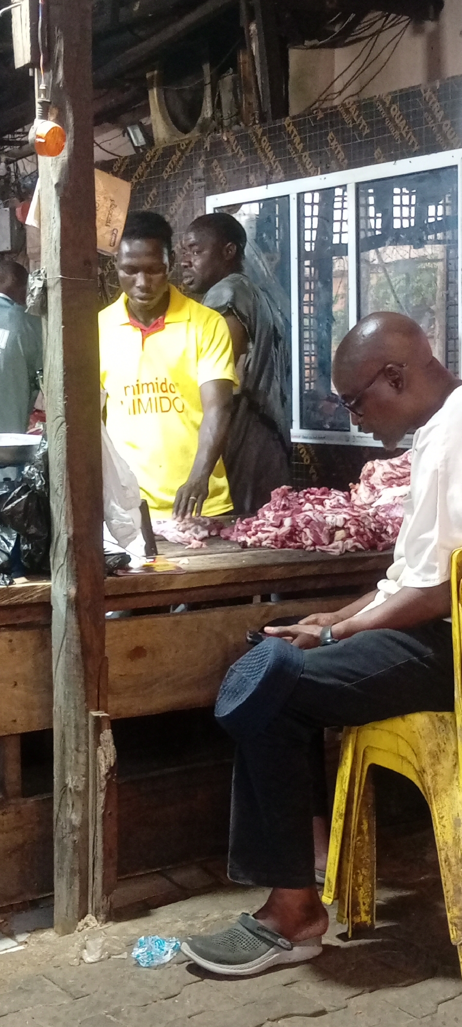 In Dorayi, Nigeria, a street-level scene depicts an open-air market stall. Two men stand behind a wooden counter laden with a substantial amount of raw meat. One man, wearing a yellow shirt with the word "nimido" printed on it, appears to be working at the stall. The second man, in a dark shirt, stands next to him. To the right of the stall, a bald man wearing glasses sits on a yellow plastic chair, looking down at a mobile phone in his hands. He is dressed in a white shirt, dark pants, and wears gray Crocs-style footwear. The stall is constructed with rough-hewn wooden posts and planks. In the background, there's a window-like structure with visible exterior elements. Cables are visible above, and general ambient lighting casts shadows throughout the scene. The ground is a paved surface with some scattered debris.