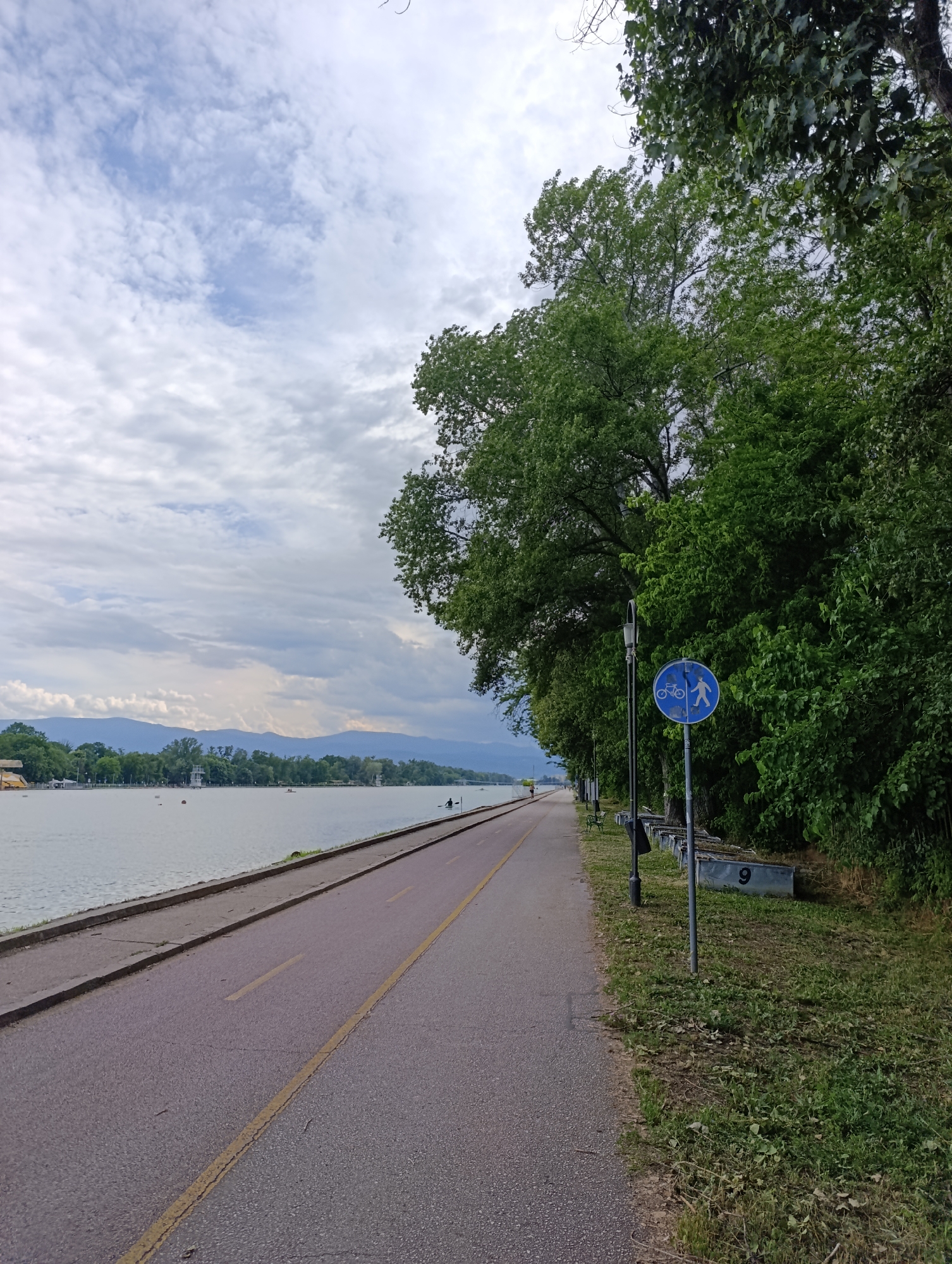 This photograph captures a tranquil waterfront scene along a paved pathway, likely the rowing canal in Plovdiv, Bulgaria. The image is dominated by a long, grey asphalt path with a clear yellow dividing line, extending into the distance.
To the left of the path, a wide, calm body of water reflects the soft, diffused light from the overcast sky, which is covered in fluffy white and grey clouds. Across the water, a distant shore reveals faint blue mountains or hills on the horizon. Several white buoys and a few small boats or kayaks, one occupied by a person, are visible on the water, suggesting recreational activities.
On the right side of the path, a dense line of vibrant green trees forms a lush natural boundary. A black, ornate streetlight stands tall beside the path. Further along, a prominent circular blue sign with white pictograms of a pedestrian and a bicycle clearly indicates a shared path for both. Below the sign, a series of grey rectangular planters lines a grassy verge, with the number "9" distinctly visible on one of them. In the far distance on the path, a lone individual can be seen walking or jogging.
The time of day appears to be daytime, under a cloudy sky, suggesting a mild and pleasant atmosphere for outdoor activities. The scene evokes a sense of peaceful recreation and exercise in a well-maintained natural setting.
