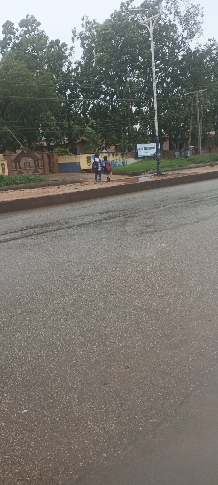 The image captures a street scene in what appears to be Bukuru, Nigeria, as indicated by the location context. Two children, likely students, are the central subjects, visible walking on a dirt sidewalk next to a paved road. Both children are wearing what seem to be school uniforms, one predominantly blue with a white backpack, and the other in a lighter-colored uniform with an orange or brown backpack.
The scene is dominated by a wide, wet-looking asphalt road in the foreground and midground, suggesting recent rainfall or damp conditions. On the left side of the road, beyond the sidewalk, there's a stone fence with an ornate gate, possibly marking the entrance to a building complex. Behind this fence, several buildings with brown roofs are partially visible, surrounded by abundant green trees and foliage, indicating a verdant environment.
A prominent street lamp with solar panels stands on the right side of the sidewalk, and further back, utility poles with power lines are also visible against the backdrop of trees and an overcast, grey sky.
A sign is clearly visible behind the children on the sidewalk, which reads "COLLEGE OF MARY IMMACULATE". This confirms the location is likely near a school or institution by that name. The overall impression is that of a cloudy day, possibly just after rain, with the children on their way to or from school.