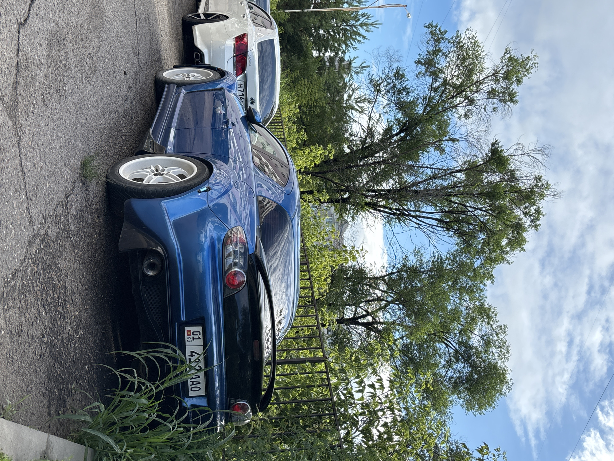 A blue sports car is parked on asphalt next to a white sedan. The sports car has a large spoiler and white rims. The license plate of the sports car reads "01 492 AAO". The white sedan has a license plate that reads "H714". In the background, there are trees and a metal fence. The sky is blue with some clouds. The image appears to be taken on a sunny day. The location is likely Blagoveshchensk, Russia, given the license plate format.