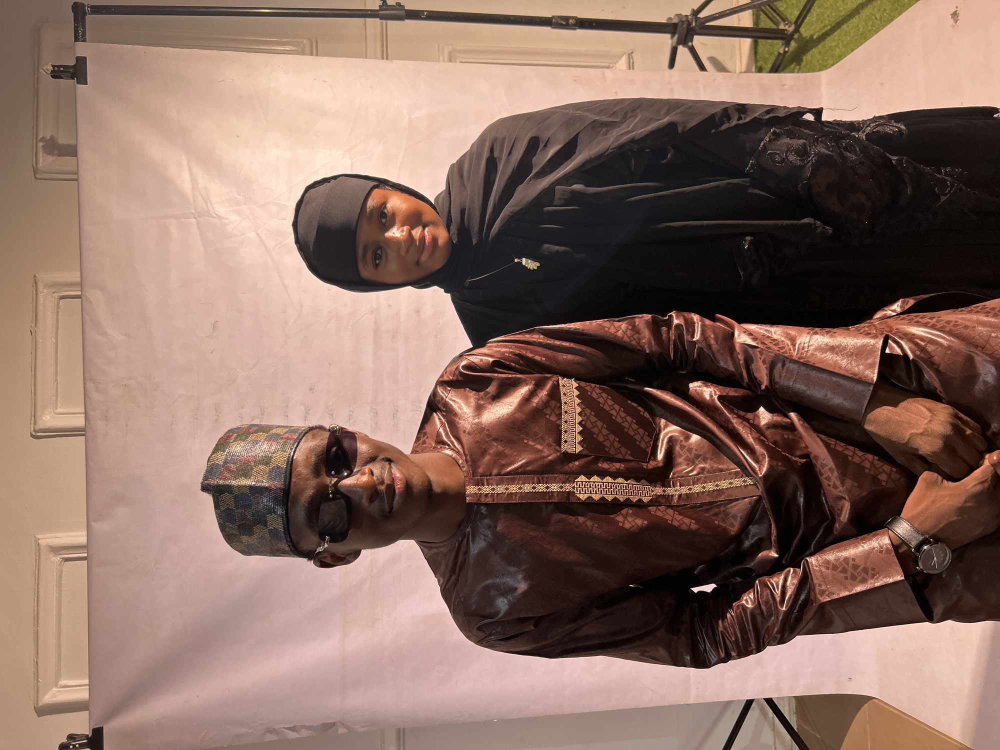A man and a woman are posing together in front of a white backdrop with lighting equipment visible. The man is wearing a patterned brown shirt and a matching hat, along with sunglasses and a watch. The woman is wearing a black hijab. The setting appears to be indoors. There is no information to suggest that this is in the city of Jos, Nigeria.