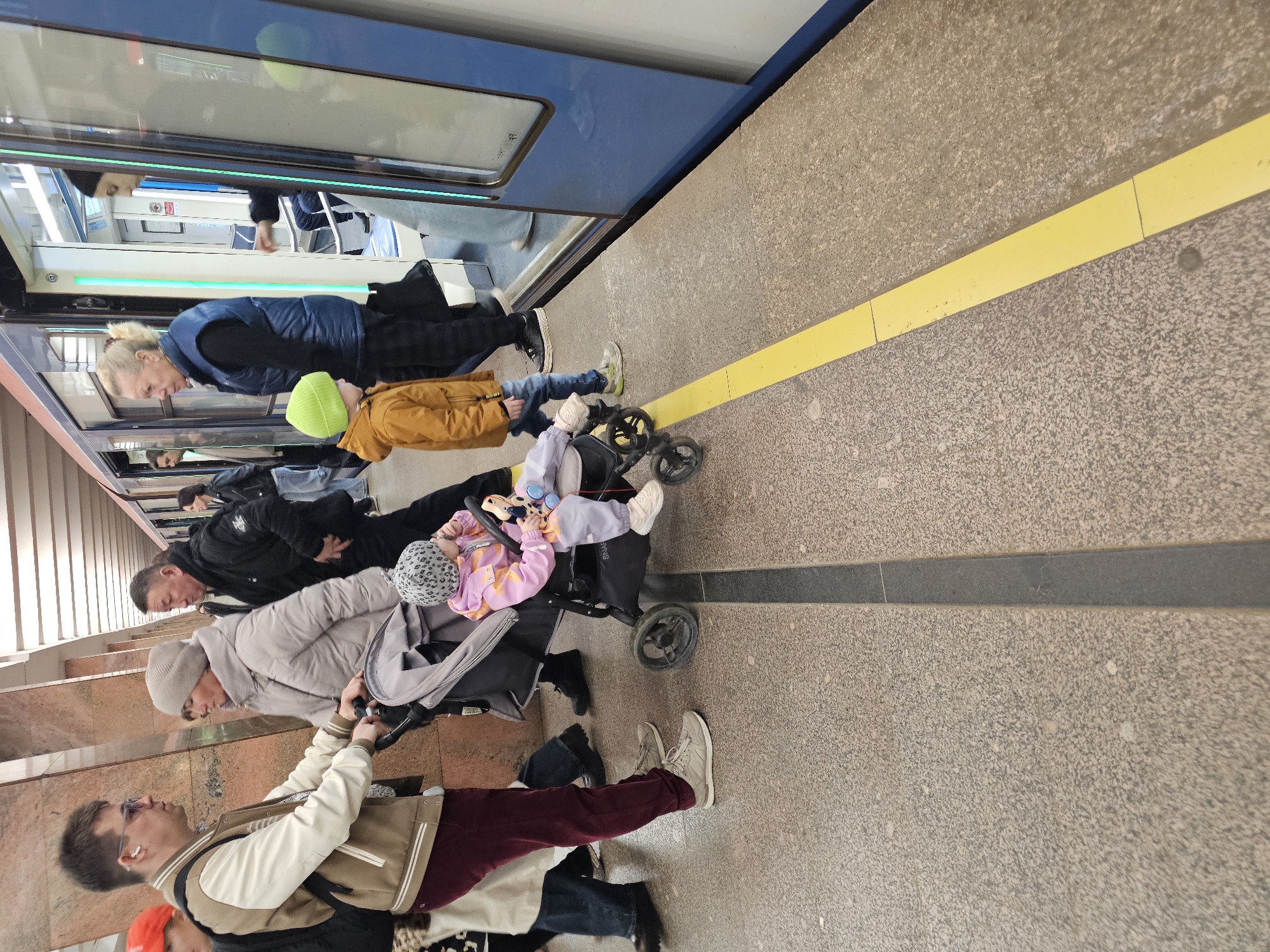The image captures a bustling platform within the Moscow Metro system, Russia. A blue metro train with open doors, illuminated by vertical green indicator lights, is stationary at the platform edge. The platform floor consists of light-colored, speckled paving with a prominent yellow safety line running parallel to the train and a dark gray line perpendicular to it.
In the foreground-left, an adult male in a brown and off-white letterman-style jacket, maroon pants, and glasses holds the handle of a gray stroller. Seated in the stroller is a toddler wearing a pink and orange jacket and a gray leopard-print beanie, holding a toy. An adult female in a light gray puffer jacket and beanie stands beside the male and stroller, facing towards the right. Behind her, another adult male in a black puffer jacket is partially visible.
To the right, closer to the train, a young child in a yellow-orange jacket, blue jeans, and a bright green beanie stands near the platform edge, observing the train. Behind this child, an adult female with blonde hair, wearing a dark blue vest, stands near the open train entrance. Another adult female is visible inside the train car through the open doors. Several other individuals are present on the platform and inside the train in the background, indicating a busy environment. Light-colored, ribbed architectural columns are visible along the platform in the background.