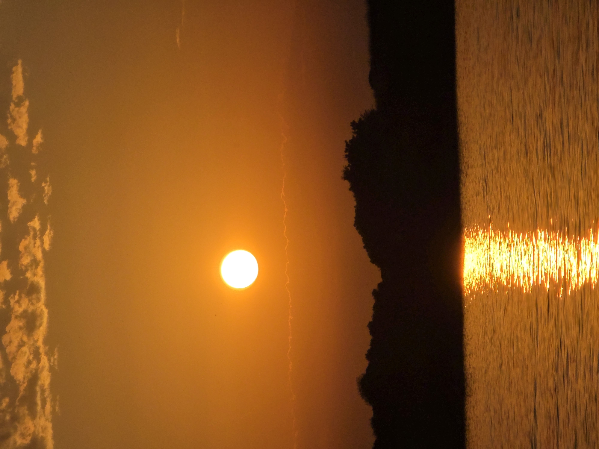 The image depicts a sunset over a body of water. The sun, a bright white orb, is low in the sky, casting a warm orange glow across the scene. Wispy clouds are scattered across the upper portion of the sky. In the foreground, the water reflects the sunset, creating a shimmering path of light. The background features a dark silhouette of trees and land. The scene appears to be a natural landscape. The location in Chernomorets, Bulgaria, is not evident from the image itself.