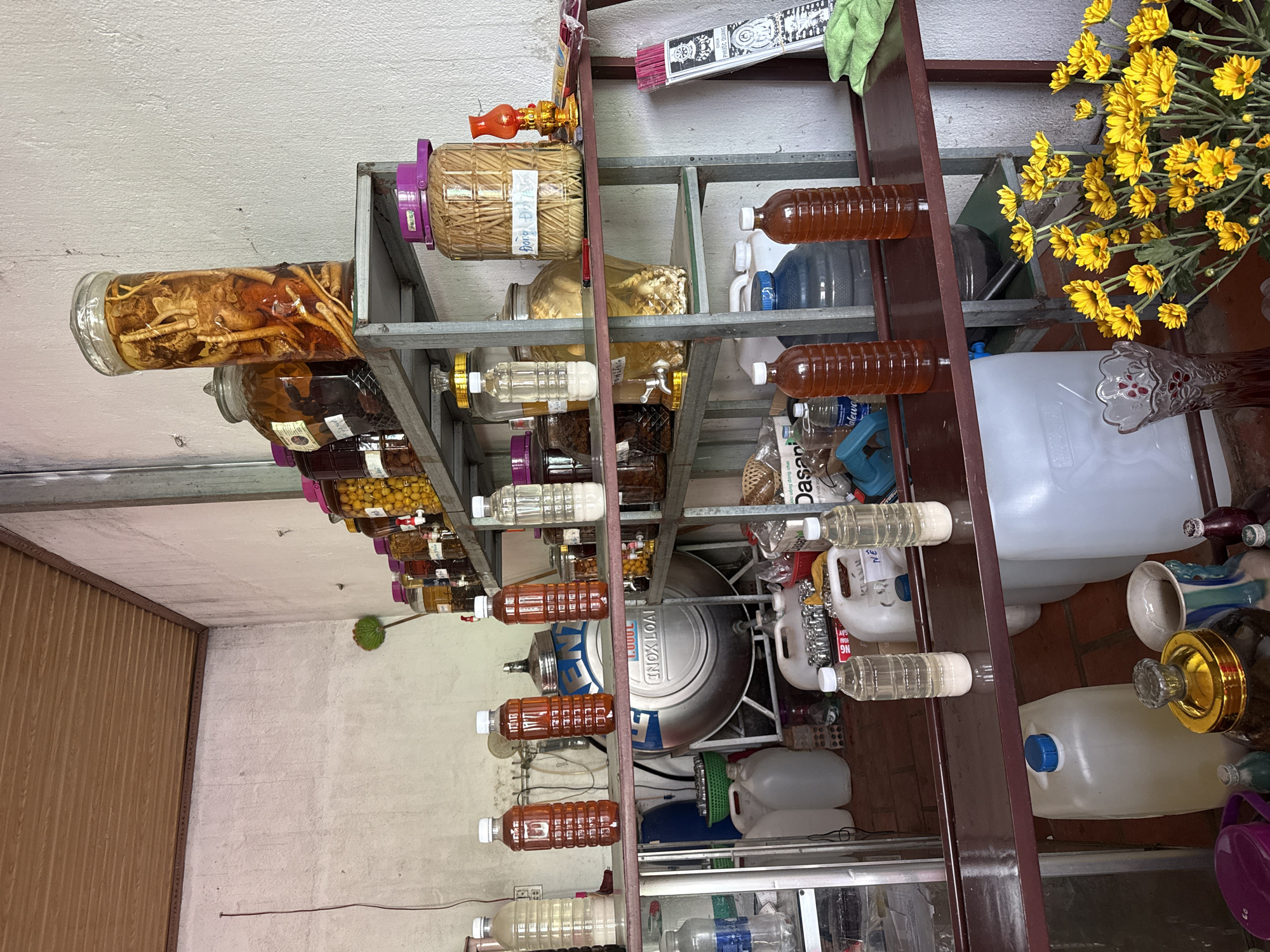 The image depicts a collection of jars and bottles filled with various liquids and ingredients, presumably for medicinal or culinary purposes, stored on metal shelves. The location appears to be Thôn 2, Vietnam, based on contextual clues. The contents of the jars range from roots and herbs to fruits and seeds, submerged in liquid, possibly alcohol or vinegar. Additional items such as water bottles, containers, and cleaning supplies are scattered around the shelves, suggesting the space is used for multiple purposes. A bouquet of yellow flowers adds a decorative touch. The overall setting is cluttered and functional.
