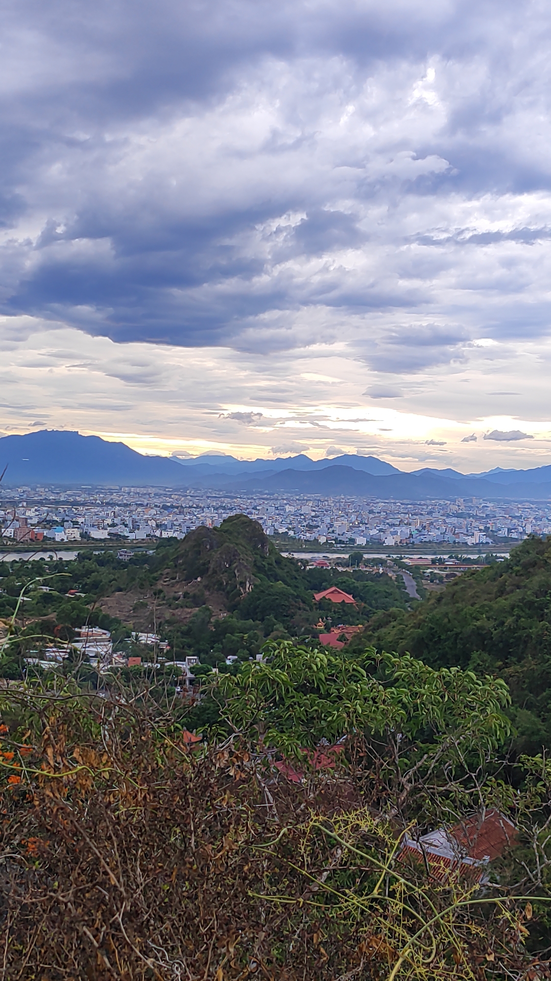This is a photograph taken from a high vantage point, looking out over a sprawling city nestled against a backdrop of mountains. The foreground is dominated by dense foliage, with dry, brown branches and green leaves creating a natural frame for the scene. In the mid-ground, a prominent, rocky hill rises from the landscape, covered in greenery. Beyond this hill, the city unfolds, a dense tapestry of buildings with various roof colors, stretching towards the horizon. The distant mountains form a rugged silhouette against a cloudy sky. The sky itself is dramatic, with heavy, dark grey clouds interspersed with patches of lighter, diffused light, suggesting either an approaching storm or the aftermath of one. The light appears to be that of late afternoon or early evening, casting a soft glow on the landscape. Given the context of Đà Nẵng, Vietnam, this viewpoint likely overlooks the city from one of the Marble Mountains (Ngũ Hành Sơn), specifically from a position on or near the Sơn Thủy beach side, looking inland. The arrangement of the city, the presence of a distinctive rock formation, and the mountainous backdrop are consistent with this location. No people are visible in the frame. The overall impression is one of natural beauty contrasting with urban development, under a dynamic sky.