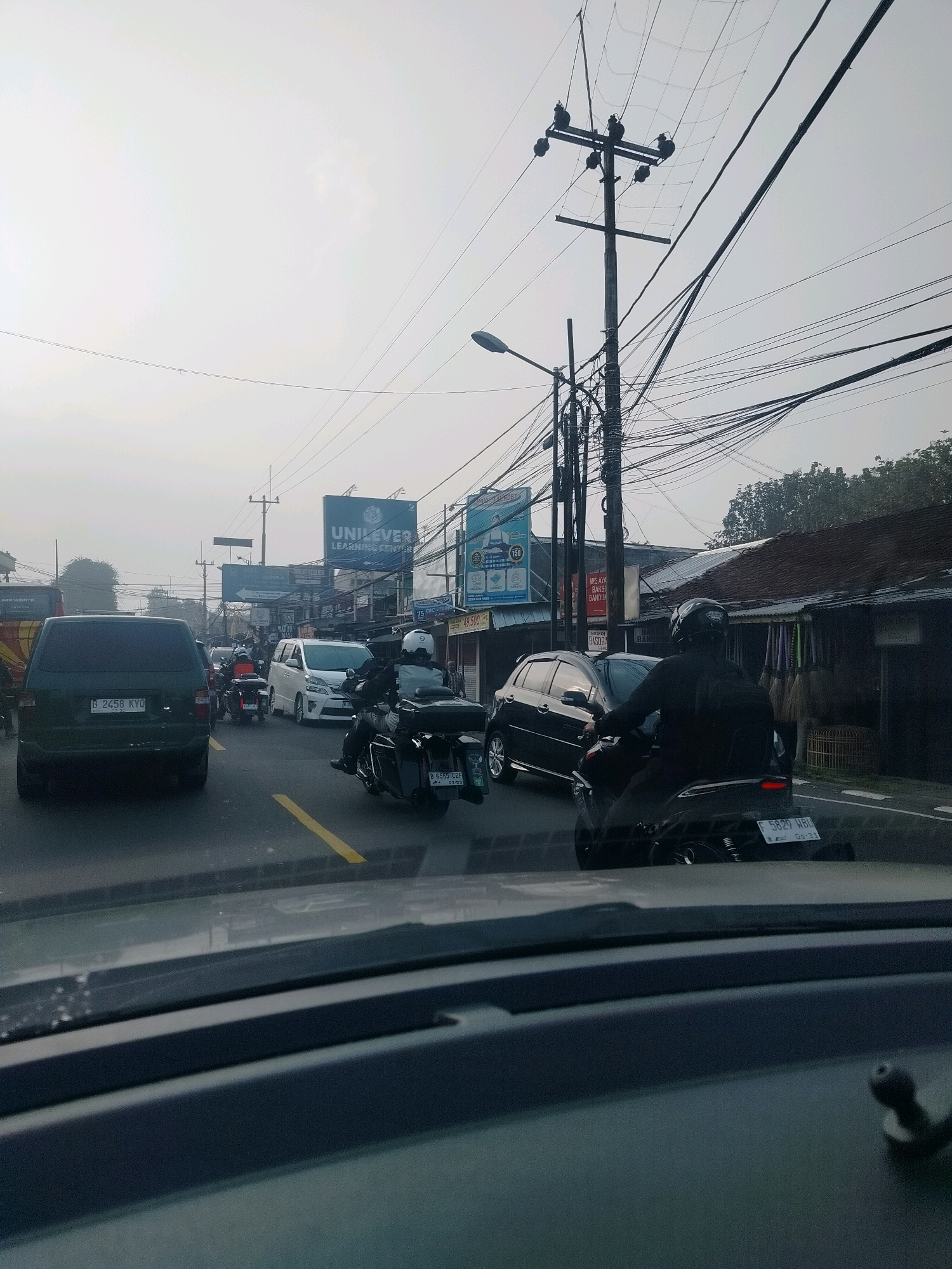The image depicts a traffic scene on a street in Megamendung, Indonesia, characterized by a moderately overcast sky. The foreground shows the interior of a vehicle, with the dashboard and windshield visible. The street is lined with various vehicles, including cars and motorcycles. A dark green SUV with license plate "B 2458 KYQ" is prominently featured in the left lane. Several motorcycles are also present, with riders wearing helmets and protective gear. In the background, there are buildings with shops and signs, including one for "UNILEVER LEARNING CENTER". Utility poles with a complex network of wires are visible overhead, indicative of urban infrastructure. The overall impression is that of a typical morning or afternoon commute in a bustling Indonesian town.