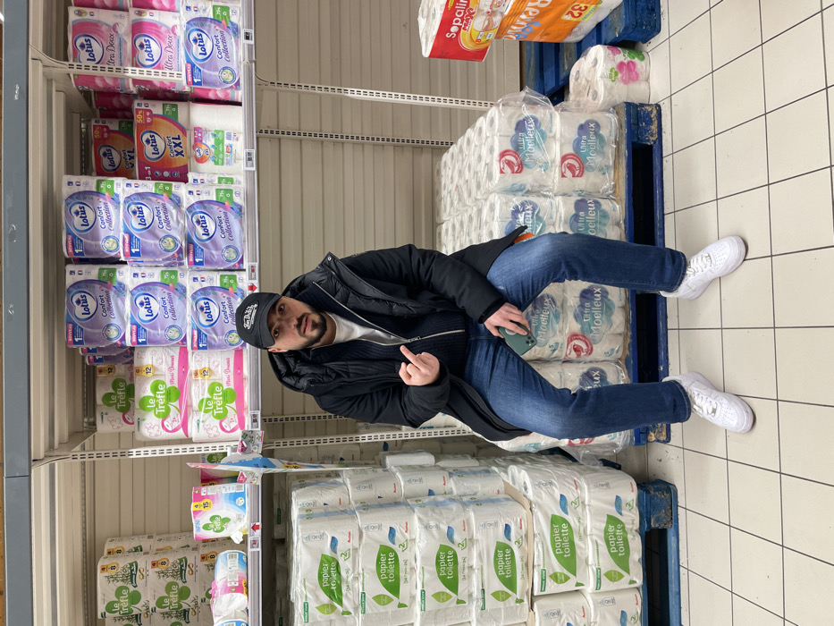 An indoor shot shows a man in a black jacket, blue jeans, and white sneakers sitting on a pallet loaded with packages of "Ultra Moelleux" toilet paper. He's in a supermarket aisle flanked by shelves stocked with various brands of toilet paper like "Lotus" and "Le Trèfle". He is holding a phone and gesturing with his middle finger raised. The floor is tiled. Overhead lighting illuminates the scene. A package of "Sopalir" toilet paper sits on top of the pallet.