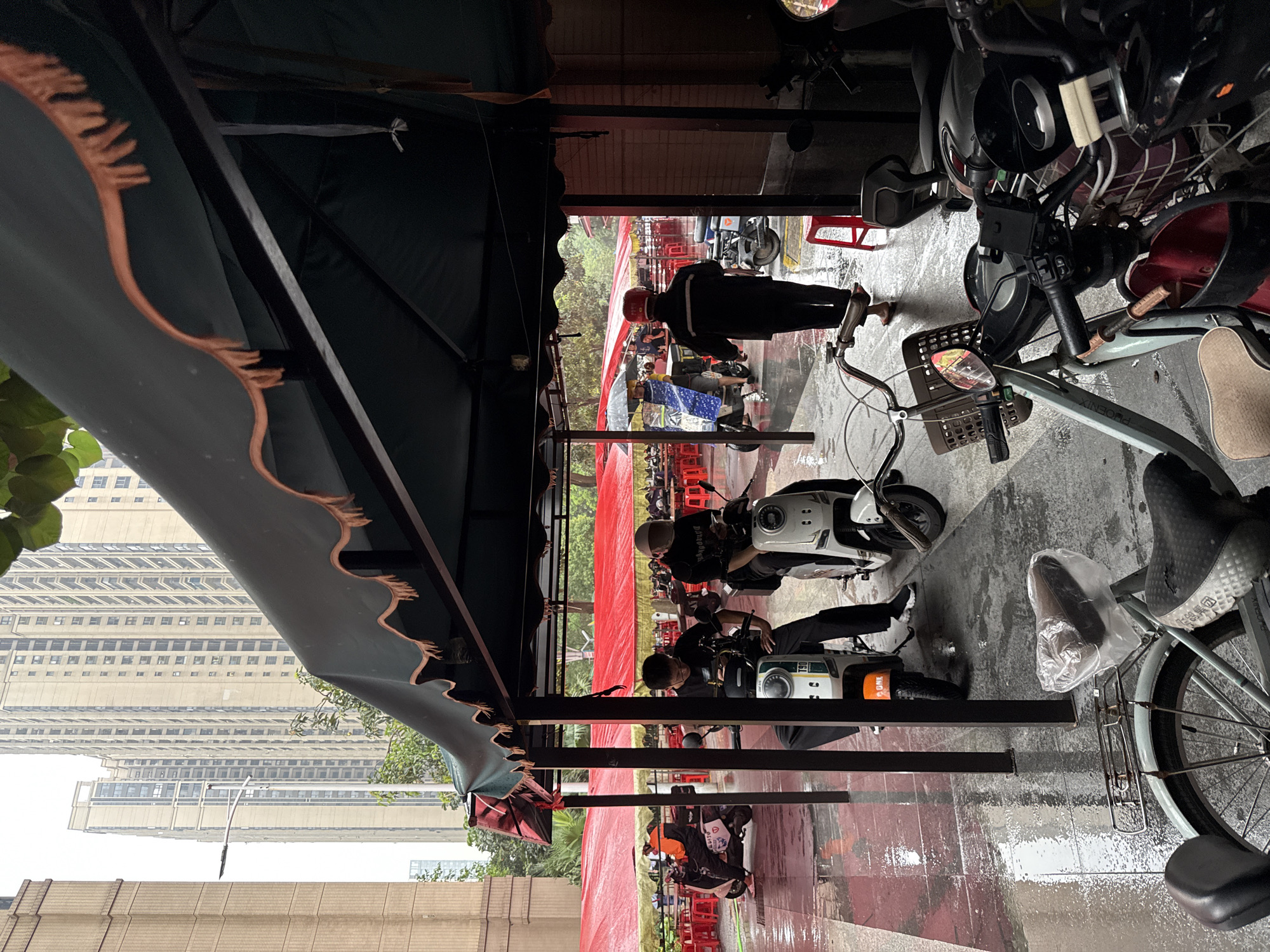 In Haizhu District, China, this image captures a rainy day scene with several people and vehicles under an awning. Two individuals are seated on electric scooters, one of whom is looking at a mobile phone. Another person, wearing a helmet and dark jacket, walks away from the camera. In the background, a line of red tables and umbrellas suggests an outdoor dining or market area. Tall apartment buildings form the backdrop of the urban environment. The wet pavement indicates recent rainfall.