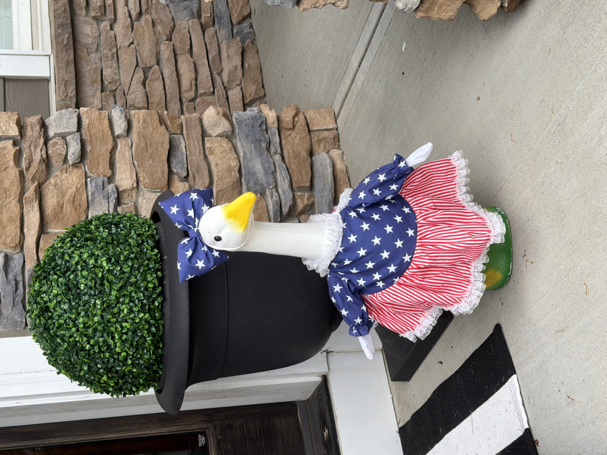 A white goose figure wearing a patriotic outfit stands on a concrete porch. The goose has a yellow beak and is adorned with a blue bow with white stars, and a dress featuring a blue star-spangled bodice and a red and white striped skirt. The dress has white lace trim. Behind the goose is a large black planter containing a round, green, leafy shrub. To the left of the goose is a black and white striped doormat. The background shows a stone-clad wall. The scene appears to be located on a residential porch.