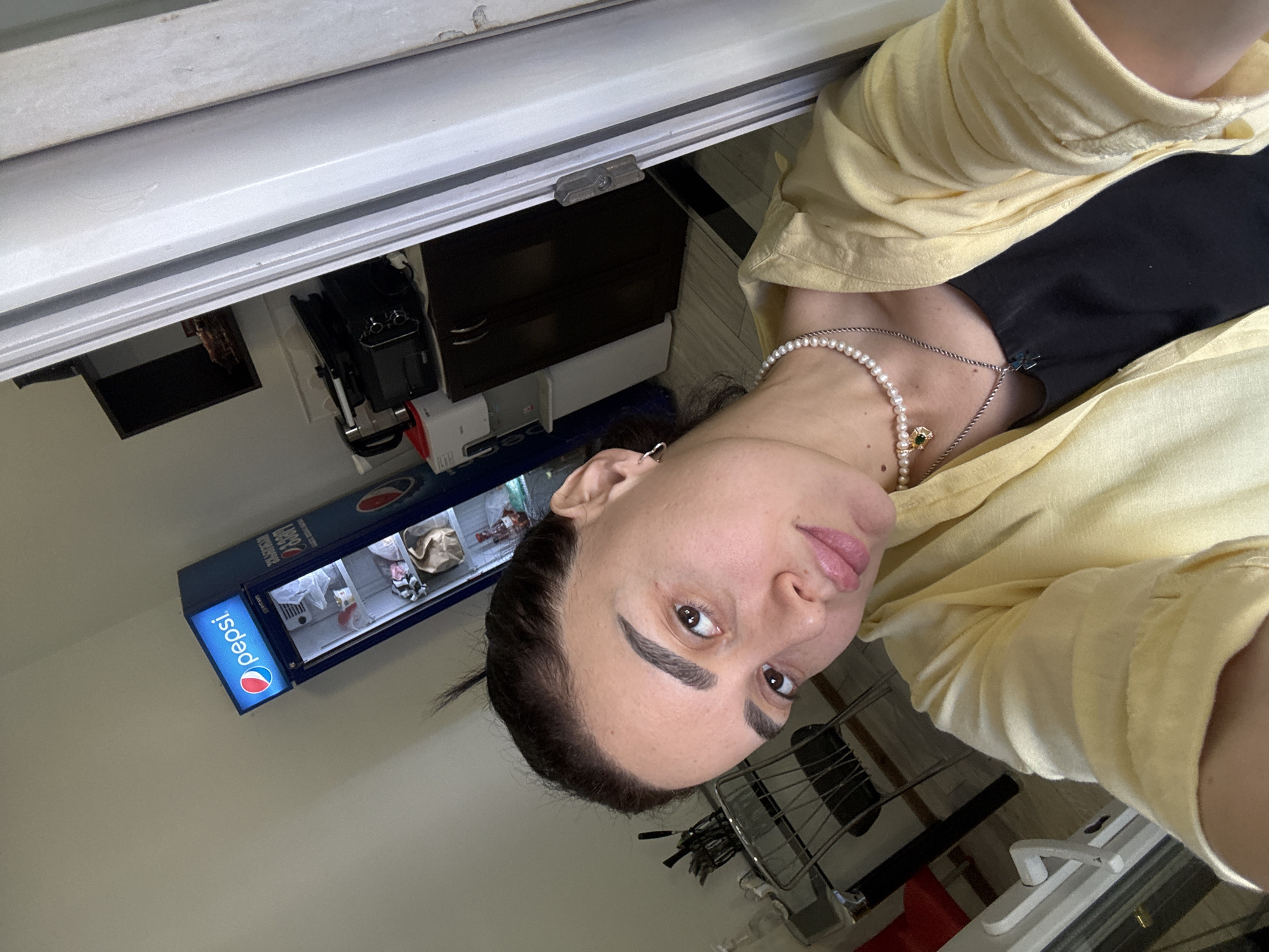 A person is looking at the camera, wearing a yellow shirt and a pearl necklace, with a black shirt underneath. The person is indoors. Behind the person is a Pepsi refrigerator and a dark brown cabinet. There is also a metal rack in the background. The location is Tbilisi, Georgia.