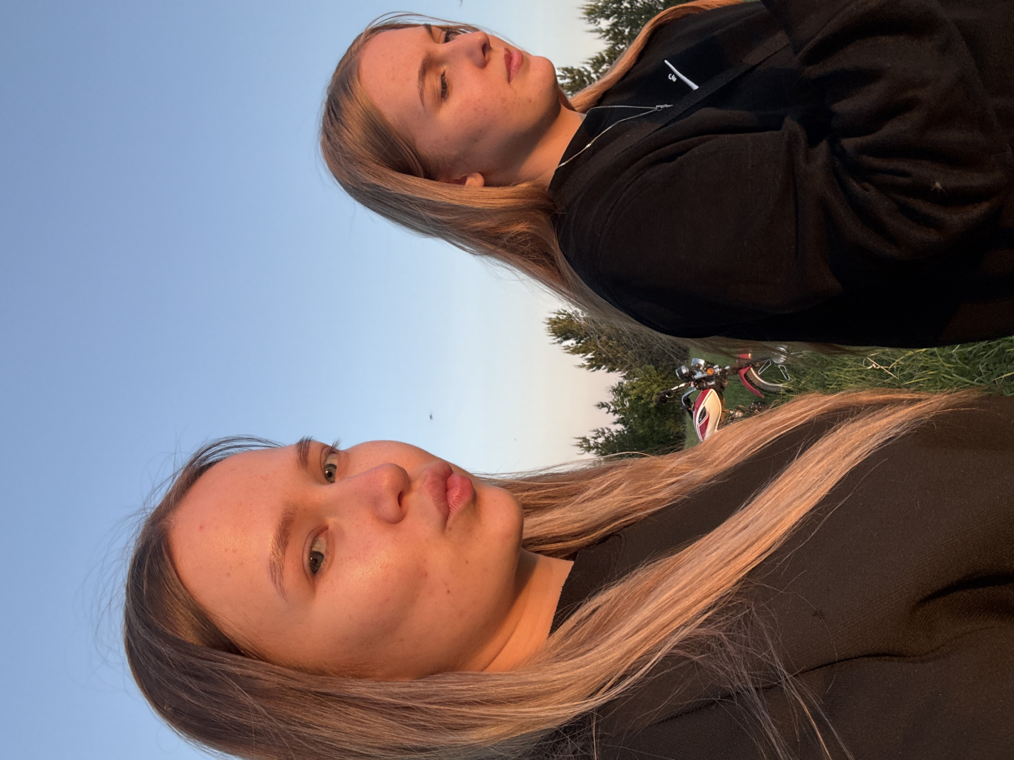 Two young women, seemingly outdoors in Zhigalki, Russia, are the primary subjects of the image. The woman in the foreground has light skin, brown/blond hair, and a slightly pouty expression. The other woman in the background also has light skin and brown/blond hair and is looking upward with her eyes mostly closed. Both women are wearing dark-colored clothing. They are on or near grass, with a bicycle partially visible in the background. The sky is light blue. It appears to be dusk or dawn.
