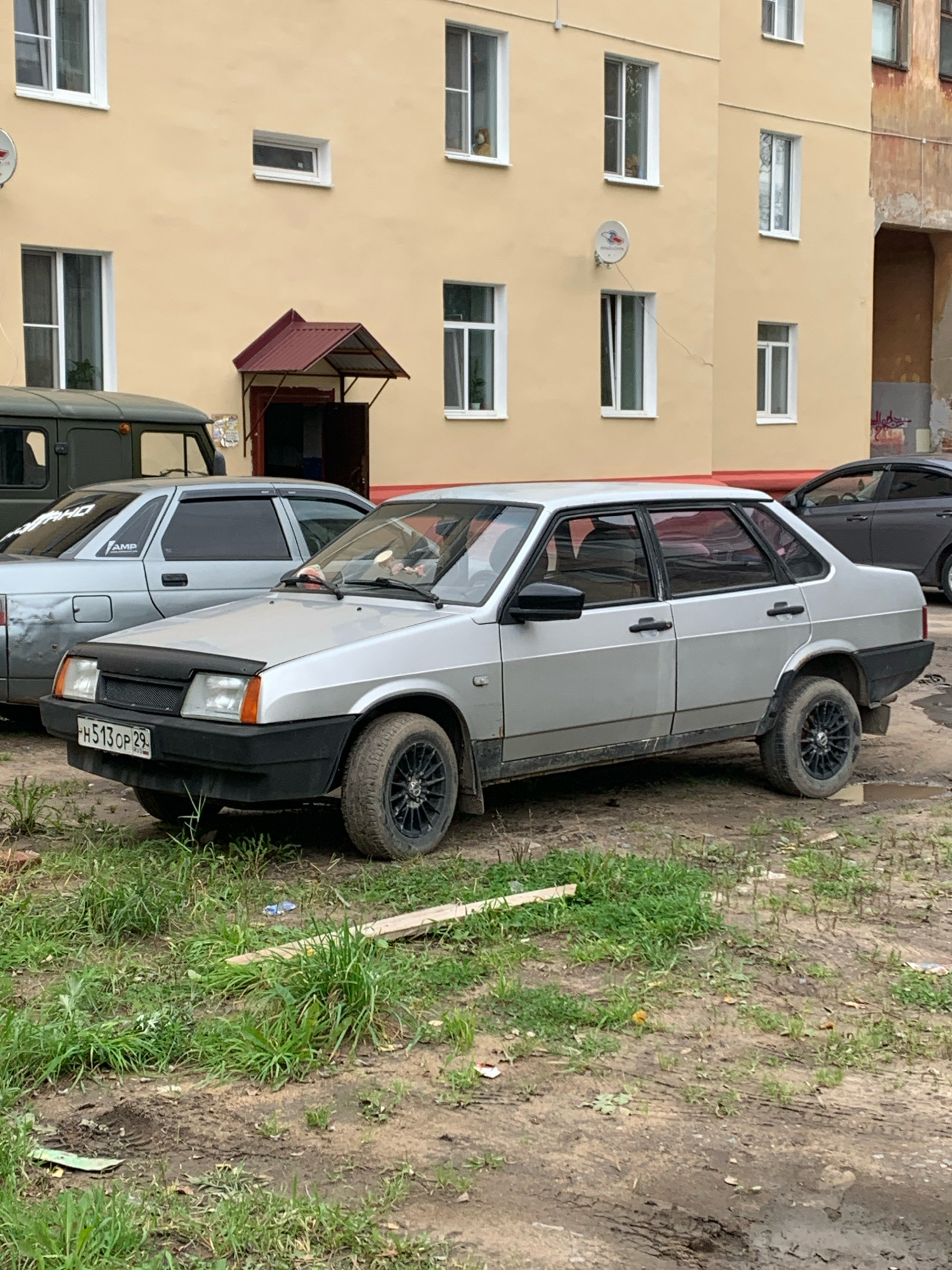 A silver Lada sedan is parked on a dirt and grass area in front of a pale yellow apartment building. The car has a black hood protector, tinted windows, and black rims. The license plate is visible and reads "H513CP 29 RUS". To the left of the Lada, the front of a dark green military-style vehicle is visible. The apartment building has several windows and a burgundy awning over a doorway. The ground is uneven with patches of dirt, grass, and some debris. The sky is not visible in the frame. The overall impression is of a utilitarian urban or suburban setting.