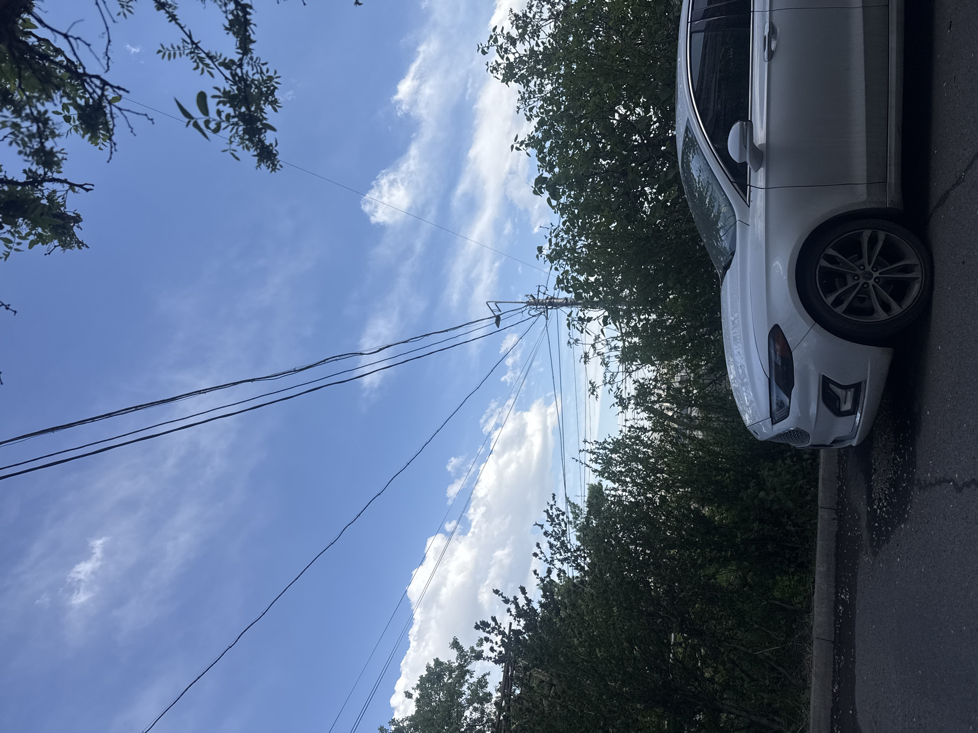 The image shows a street scene in Yerevan, Armenia. A white car is parked on the right side of the frame, adjacent to a sidewalk. Overhead, power lines stretch across a blue sky with scattered clouds. Trees with green foliage border the scene. The asphalt street has some visible wear.