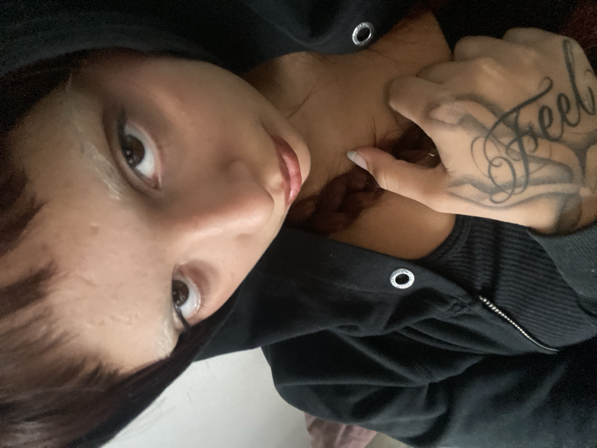 A close-up, angled shot depicts a woman with brown hair and eyes. She is wearing a black hooded sweatshirt that is partially zipped. Her hand, adorned with a "Feel" tattoo, is near her neck. No details suggest a specific location in Buenos Aires, Argentina.