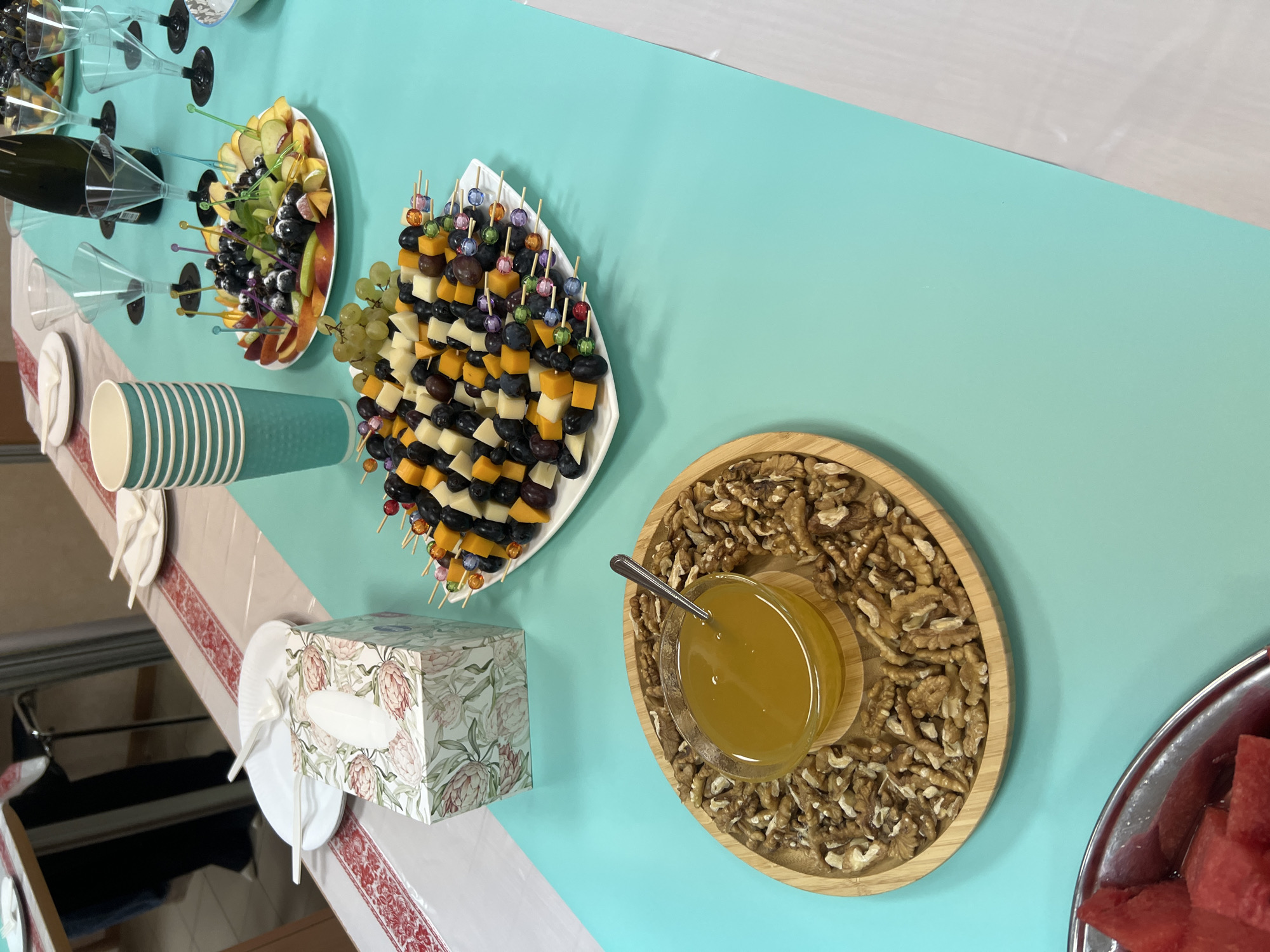 This is a forensic, matter-of-fact description of a table setting. The image shows a turquoise table covered with various items. On the left, there's a wooden platter with whole walnuts surrounding a small glass bowl of honey, which has a spoon in it. To the right of the walnuts and honey is a white rectangular platter with skewers of fruit and cheese. The skewers contain cubes of cheese, grapes, blueberries, and what appear to be pieces of fruit like melon or peach. In the background, there are several clear plastic wine glasses, some empty and some appearing to have dark liquid in them, next to a bottle of dark liquid. There are also teal-colored paper cups stacked on the left side of the table. A box of floral-patterned tissues is also visible. The table is set in what appears to be an indoor event space, possibly a home or small venue. The image is taken from a slightly elevated, top-down perspective. There are no discernible individuals in the immediate foreground or midground actively interacting with the items. The environment appears to be indoors, with a light-colored wall visible in the upper left background and a patterned tablecloth on a table in the far background. No specific information or visual cues suggest the location is Volgograd, Russia.
