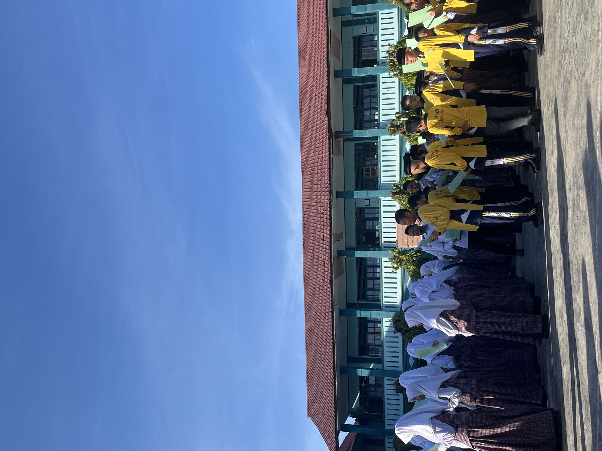 This image depicts a group of students standing in front of a teal-colored school building with a red tiled roof. The students are a mix of boys and girls, with the girls wearing white hijabs and patterned skirts, and the boys wearing yellow shirts with navy blue accents and caps. Some students are holding green papers. The scene appears to be outdoors, likely on school grounds, with a clear blue sky overhead. The architectural style of the building suggests it is located in Indonesia. Based on the context of the students' attire and the building's appearance, this could be a school in the city of Lhokseumawe, Indonesia, possibly during a school event or assembly. The lighting indicates it is daytime, with shadows cast on the ground.