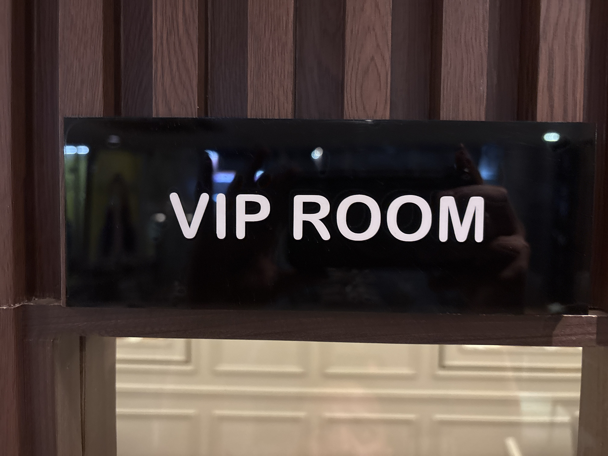 The image shows a black rectangular sign with "VIP ROOM" written in white sans-serif font. The sign is mounted on a wooden vertical panel wall. Below the sign, a portion of a glass door or window is visible, revealing a light-colored interior with a pattern of horizontal and vertical lines. A reflection is visible on the black sign, showing a person's head and shoulders, and some lights. The scene appears to be an entrance to a private or exclusive area, possibly a lounge, club, or hotel, likely located in Pekanbaru, Indonesia, given the context.