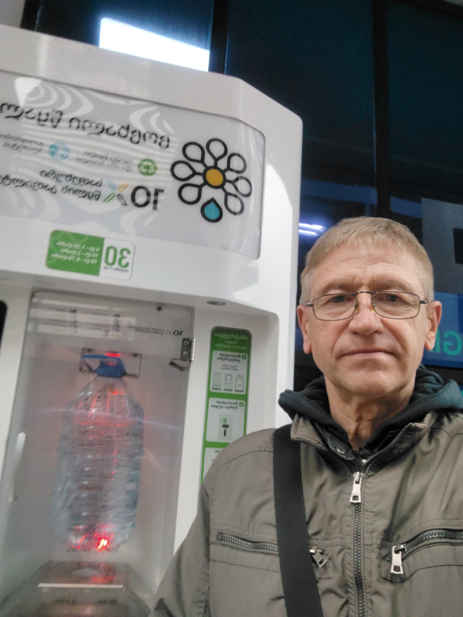 An older, light-haired man with glasses stands next to a water refilling station in what appears to be Batumi, Georgia. He is wearing a grey jacket over a hooded sweatshirt and a black strap is slung across his chest. The refilling station is white with a visible plastic bottle inside a compartment, illuminated by red light. Various symbols and text in Georgian script are visible on the machine, including the letters "OR" in Latin script. The background shows what appears to be the interior of a building with window panels.