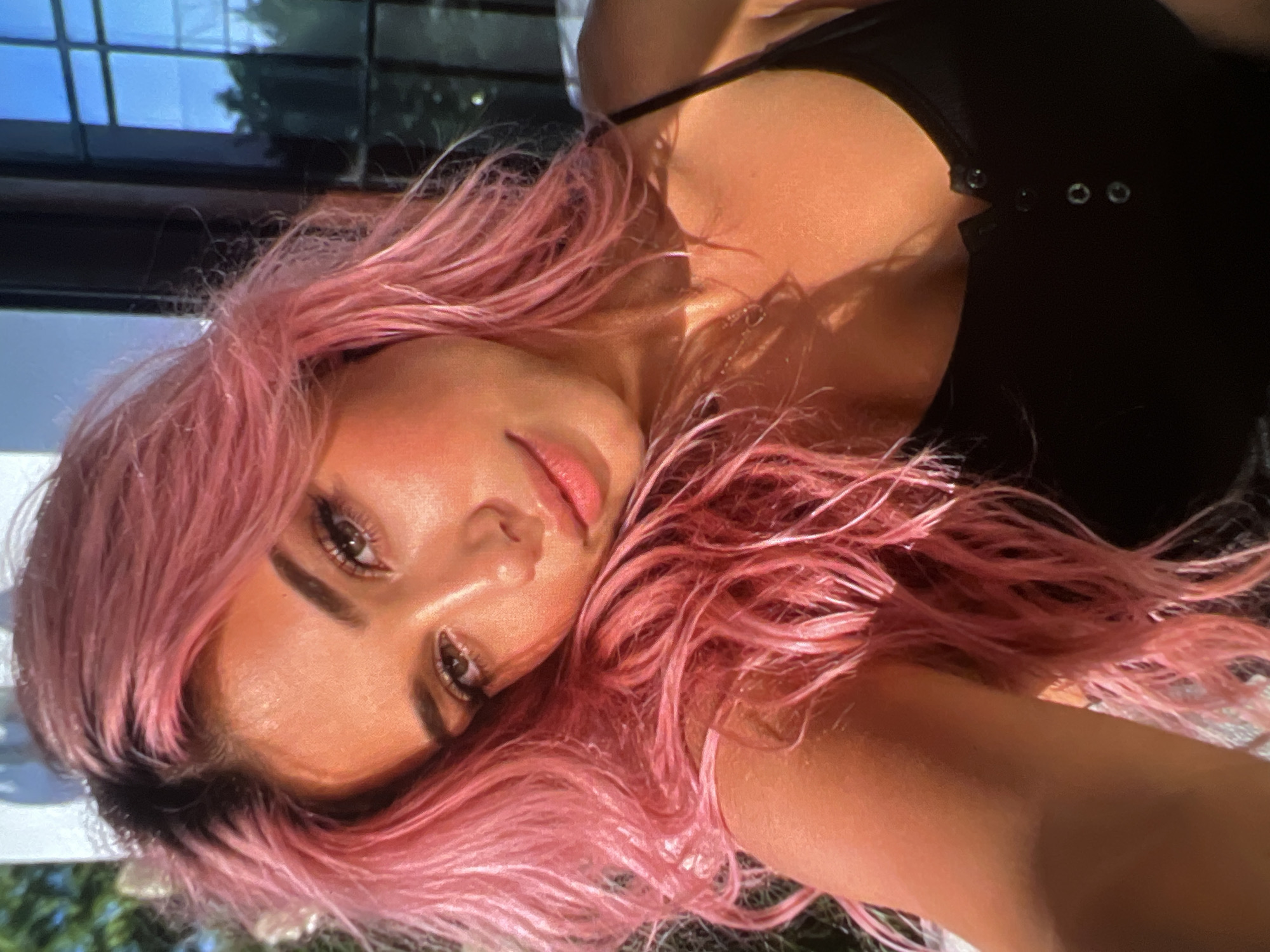 A woman with bright pink hair takes a selfie. She is wearing a black tank top. Her face is illuminated by warm, golden light, and her hair frames her face. She appears to be outdoors, with some greenery visible in the background. A window with reflections can be seen to her right.