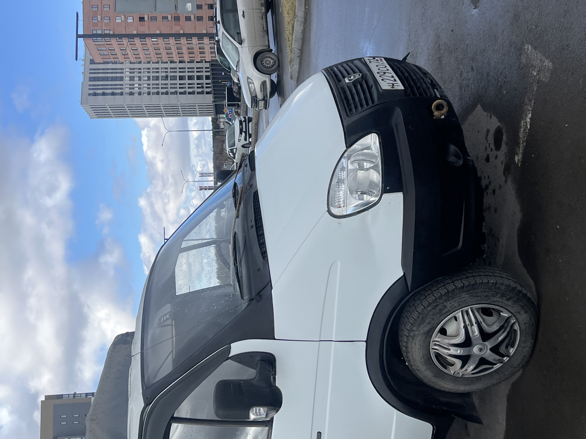 The image shows a white GAZelle van parked on a wet city street, likely in Tyumen, Russia, based on the vehicle's license plate (H229CO72). The van is the primary subject, with focus on its front passenger side, including the front headlight, grill, and wheel. In the background, a few other parked vehicles can be seen, and behind them, tall apartment buildings, reflecting the urban setting. The sky is visible above the buildings, showing a mix of blue sky and clouds. The ground is wet, reflecting the scene. The overall scene suggests an overcast day in an urban location in Tyumen, Russia.