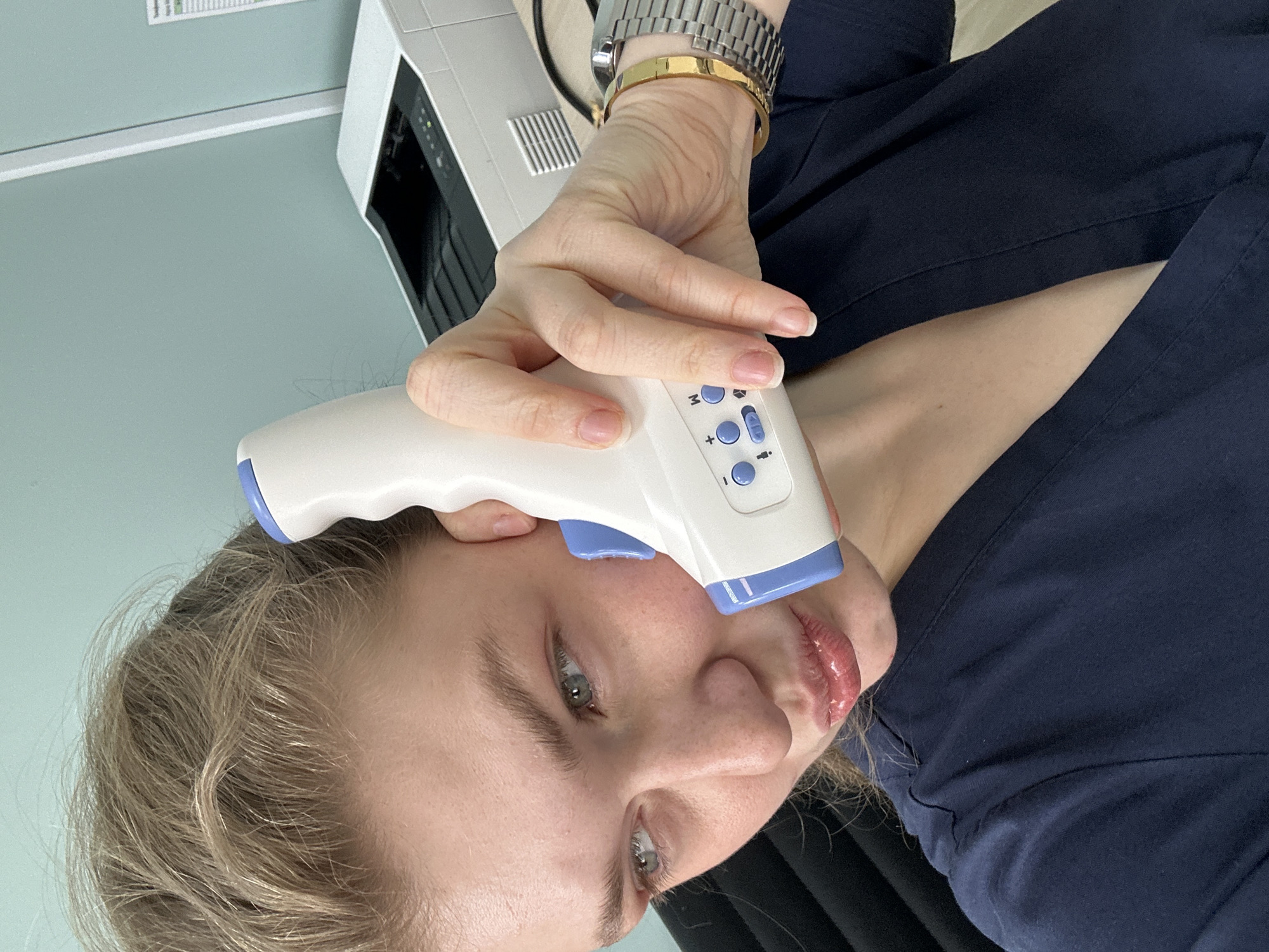 A woman is holding a non-contact forehead thermometer to her forehead to take her temperature. She is wearing a silver watch and a gold bracelet on her left wrist, and a dark blue shirt or uniform. The background includes a light blue wall and a white printer. The location cannot be confirmed as Kazan, Russia, from the image alone.