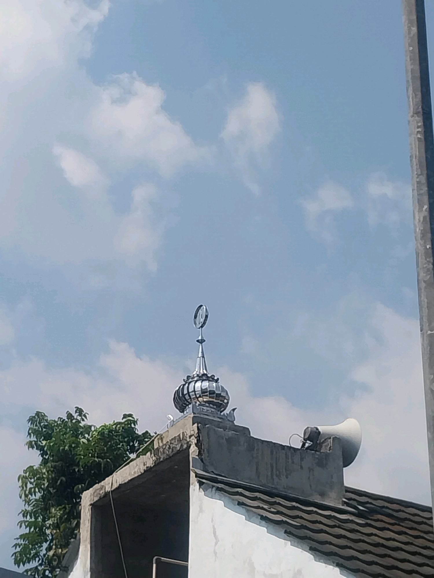 The image shows the top of a building, possibly a mosque, in Sleman, Indonesia. The building has a tiled roof and a dome with a crescent moon emblem. A speaker is mounted on the roof. A tree with green leaves is visible to the left of the building. The sky is blue with scattered clouds. A vertical pole or pillar is visible on the right side of the image. The scene appears to be outdoors during the daytime.