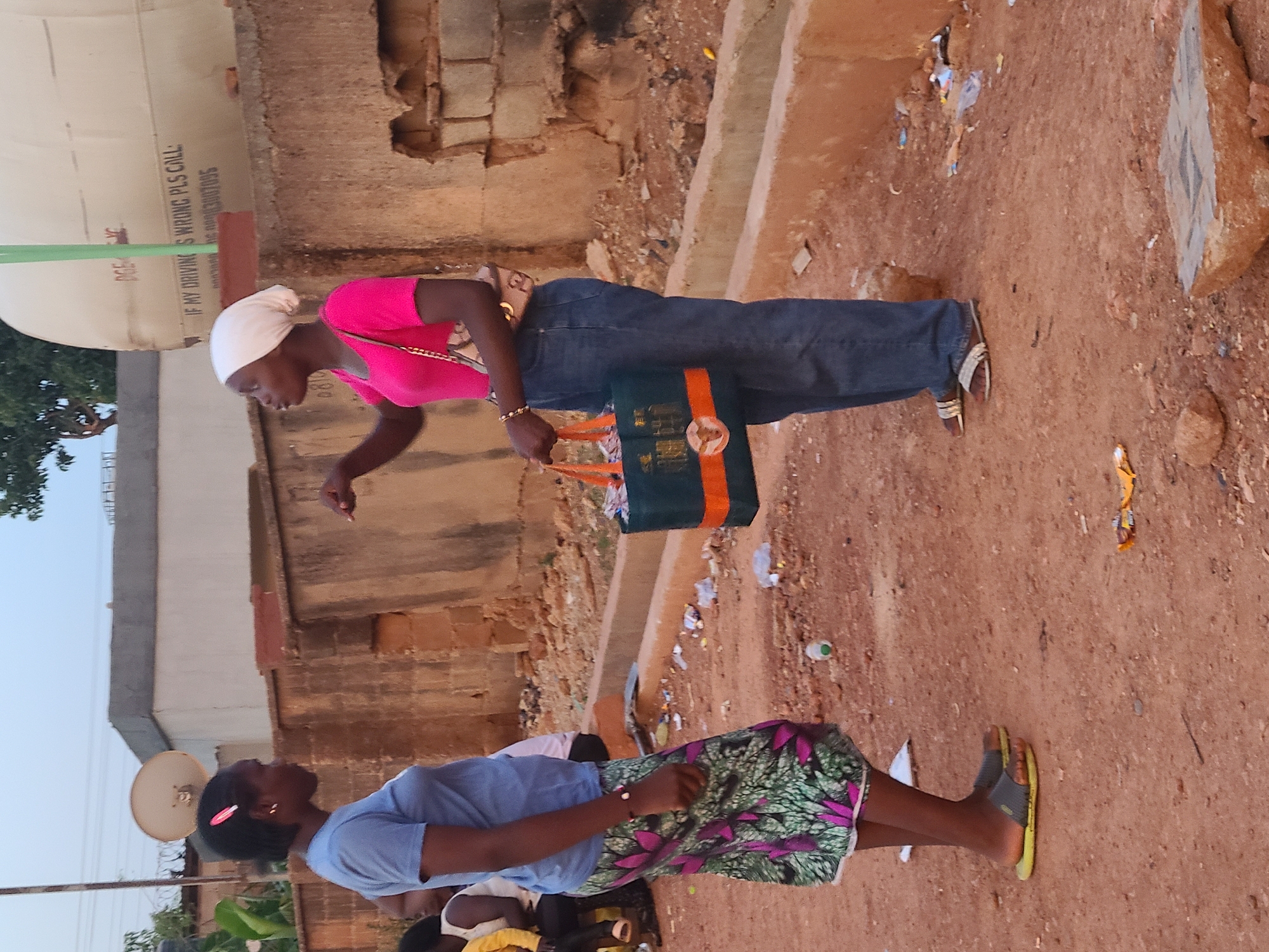 Two women are standing in an outdoor area with a dirt ground. One woman wears a pink top, blue jeans, a white head covering, and sandals. She is holding a green bag with orange stripes and pointing with her right hand. The other woman wears a light blue top and a colorful skirt, and sandals. Buildings and a water tank are visible in the background. Debris is scattered on the ground.