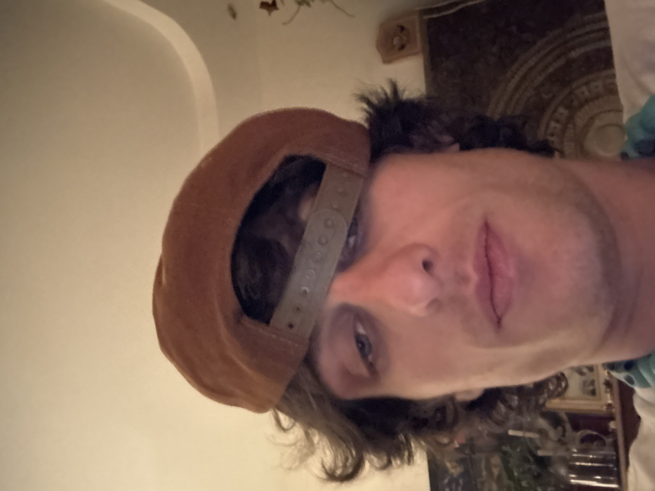 A person wears a tan baseball cap backwards, revealing a glimpse of dark hair. The individual faces the camera with light skin, a prominent nose, and lips slightly parted. Their eyes are partially visible. The background includes a cream-colored wall on the left and a framed object with an ornate design on the right.
