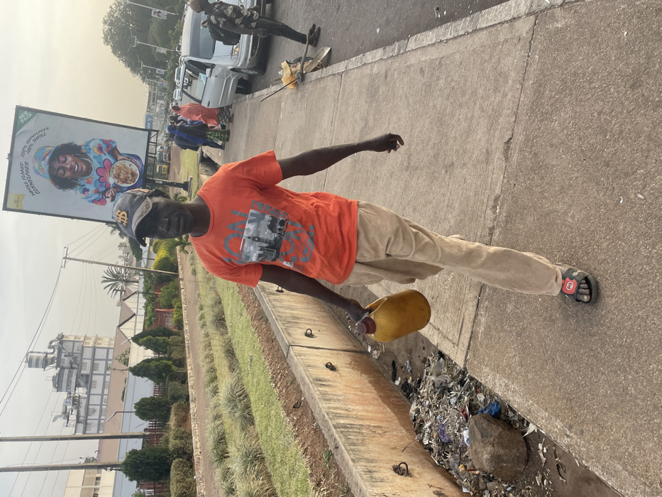 A man in an orange shirt and khaki pants holds a yellow plastic container, pouring liquid into a roadside drain filled with trash. He wears a baseball cap and sandals. A billboard with a smiling woman and food stands in the background, along with buildings and a few parked cars. The scene is an urban environment on a sunny day.