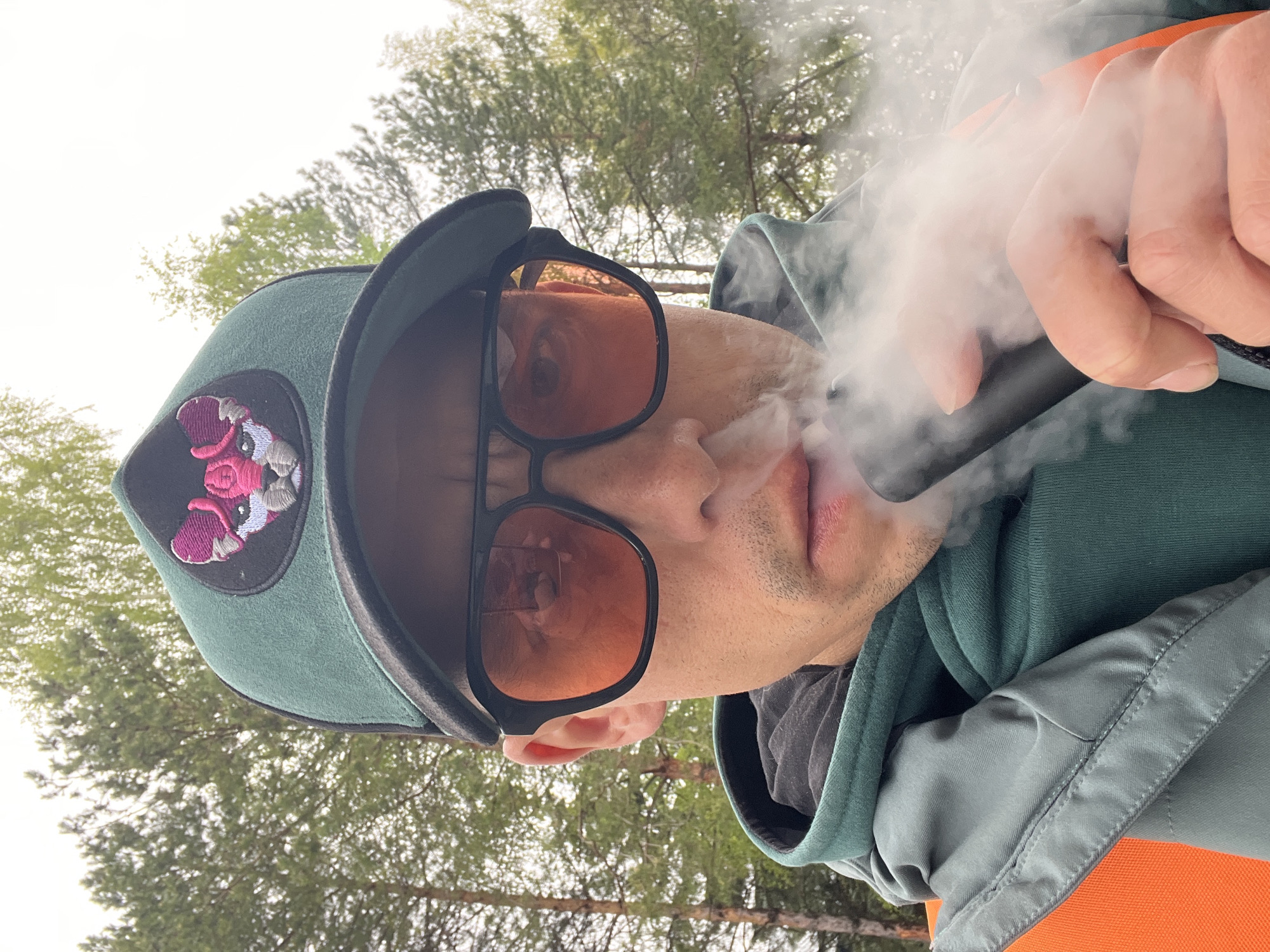 A close-up shot depicts a person vaping. The individual is wearing a green cap with a patch featuring a pink and white design, sunglasses with orange lenses, and a green hooded sweatshirt. The person is holding a black vaping device, and a cloud of vapor is emanating from their mouth. The background reveals a view of trees. While the image does not confirm, the request suggests this scene may have been taken in Chaikovsky, Russia.