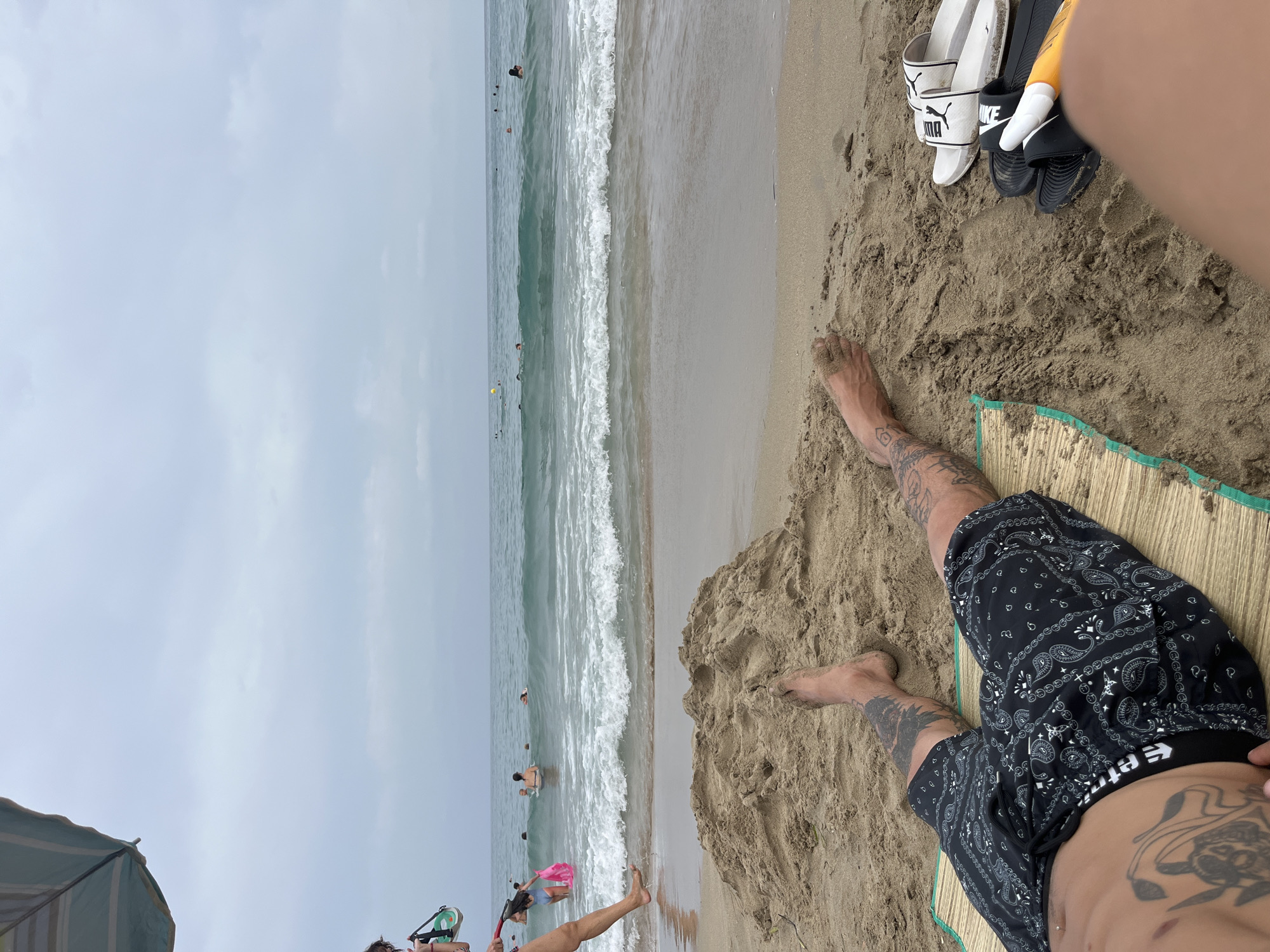 A first-person perspective shot captures a serene beach scene in Urbanització Venta De Lanussa, Spain. The foreground features the legs and torso of a person lying on a woven mat, adorned with extensive tattoos. A pile of sand is visible to the left, and a pair of slides and a sunblock tube rest on the sand to the right. Beyond the sandy shore, the tranquil sea stretches towards the horizon under an overcast sky. Several people are scattered across the water, some swimming and others wading near the shore. A blue and green striped umbrella is partially visible in the upper left corner, suggesting a shaded relaxation spot. The overall atmosphere is one of peaceful leisure.