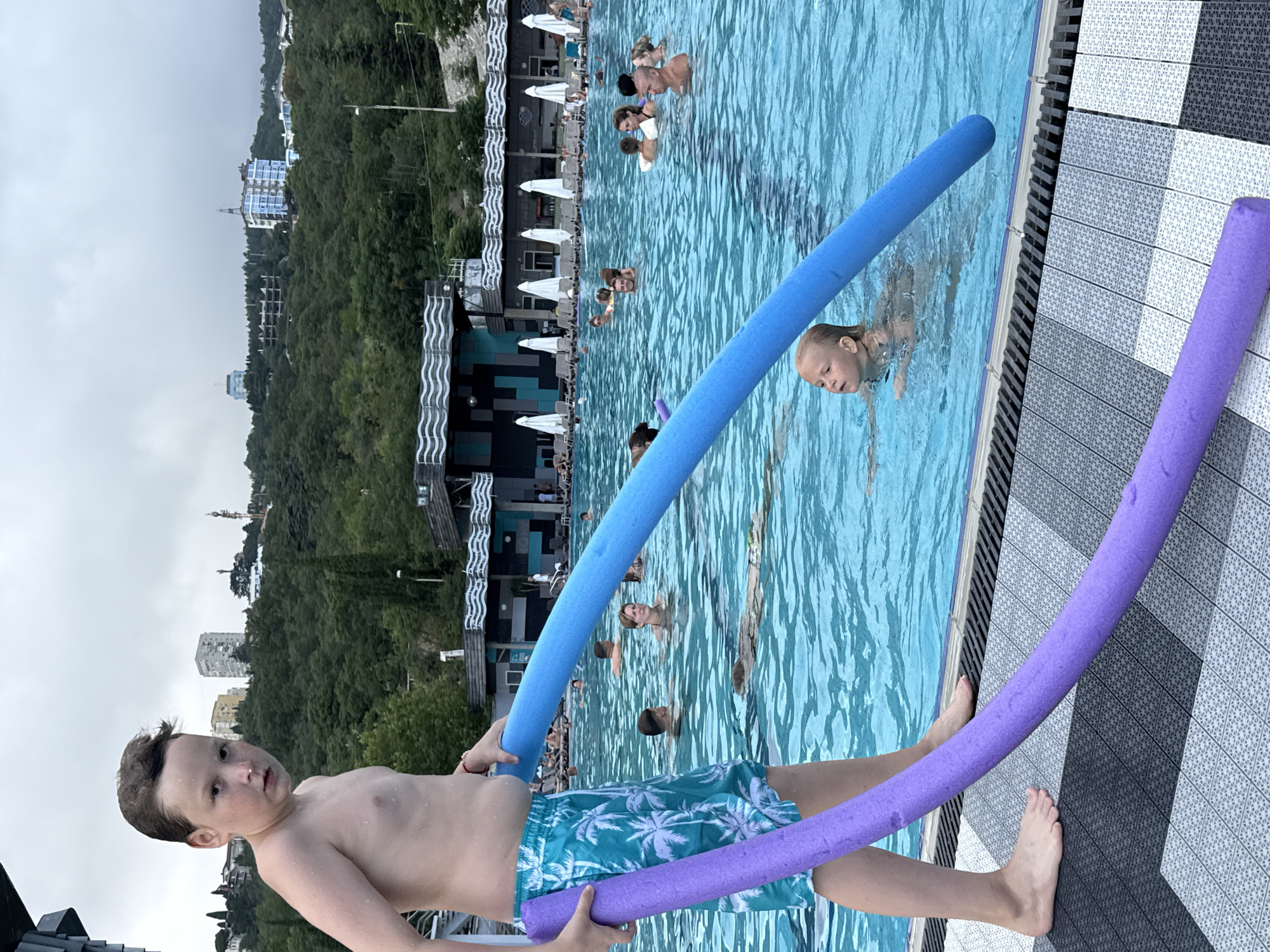 The image shows a boy standing on the edge of a swimming pool, holding two pool noodles, one blue and one purple. He is wearing blue and green patterned swim trunks. The pool is filled with people, some of whom are swimming and others are relaxing on lounge chairs in the background. The setting appears to be a resort or hotel with buildings and lush green trees on a hillside in the distance. The sky is overcast, suggesting a cloudy day. The overall atmosphere seems relaxed and recreational, with people enjoying a swim. The pool noodles are a prominent object in the foreground, suggesting playfulness or preparation for water activities.